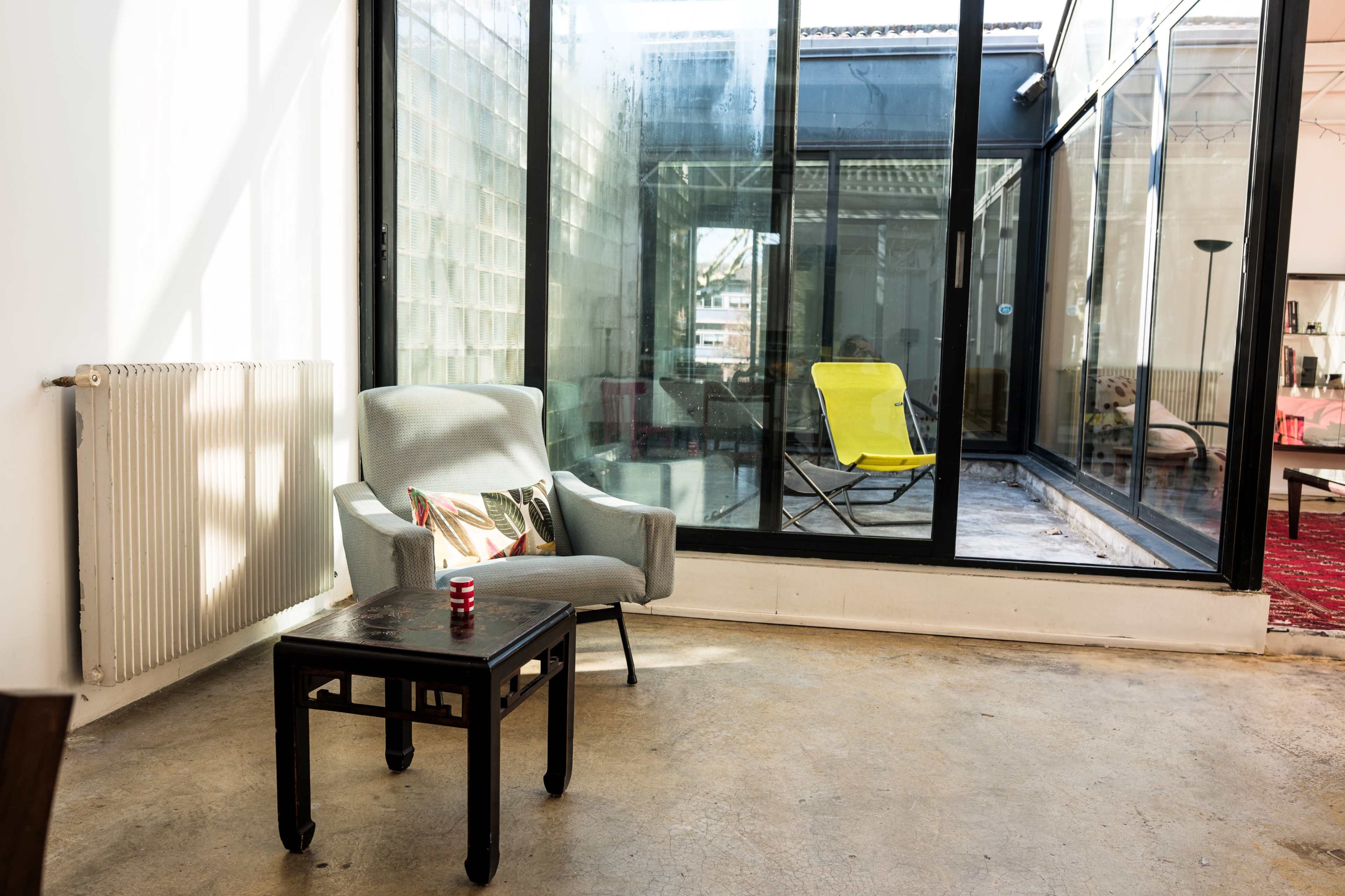 A gray armchair and a black side table are positioned near a large glass window overlooking an enclosed outdoor area.