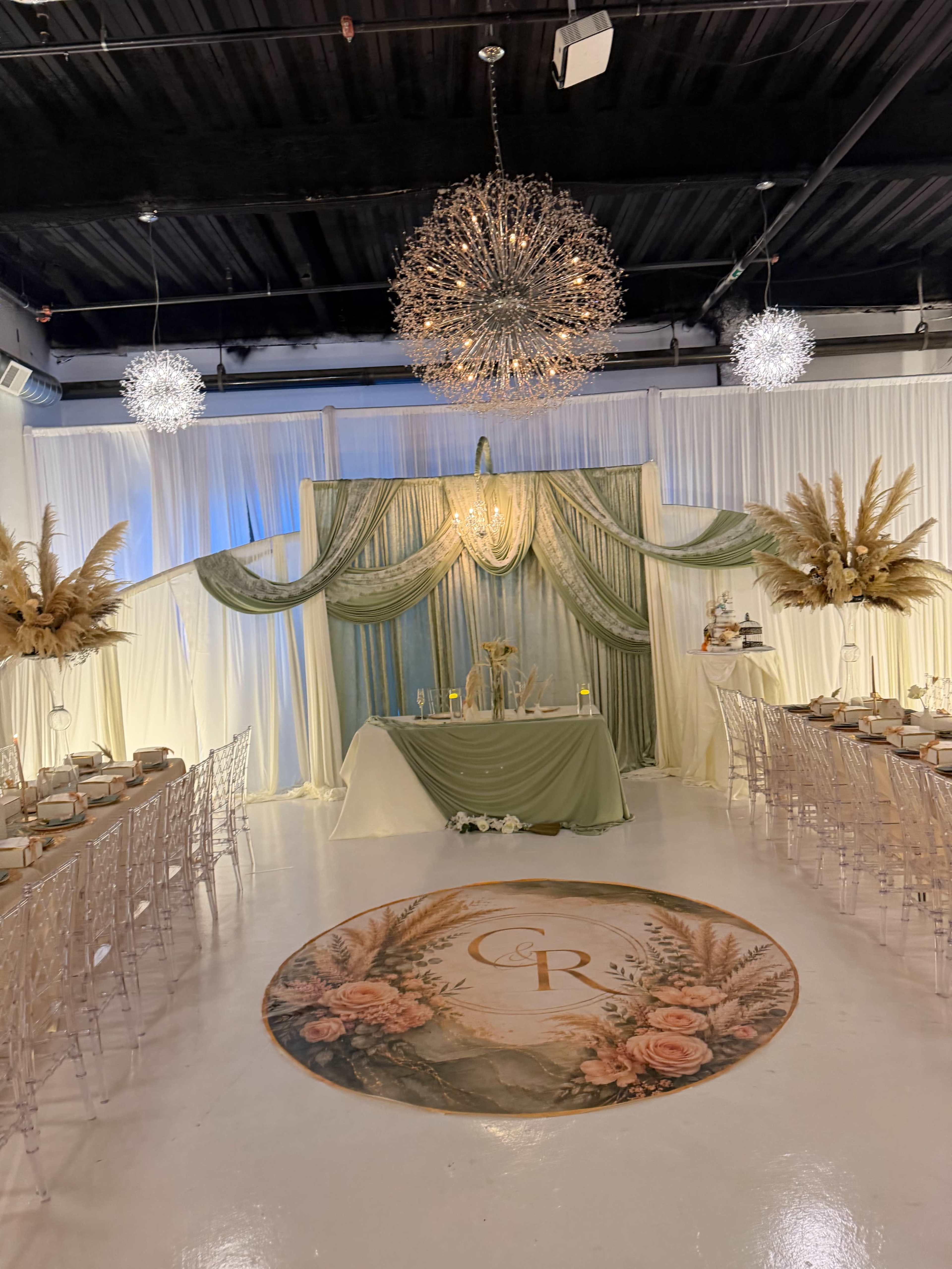 The image shows an elegantly decorated banquet hall with a central backdrop draped in fabric, adorned with decorative elements and a large round floral design on the floor.
