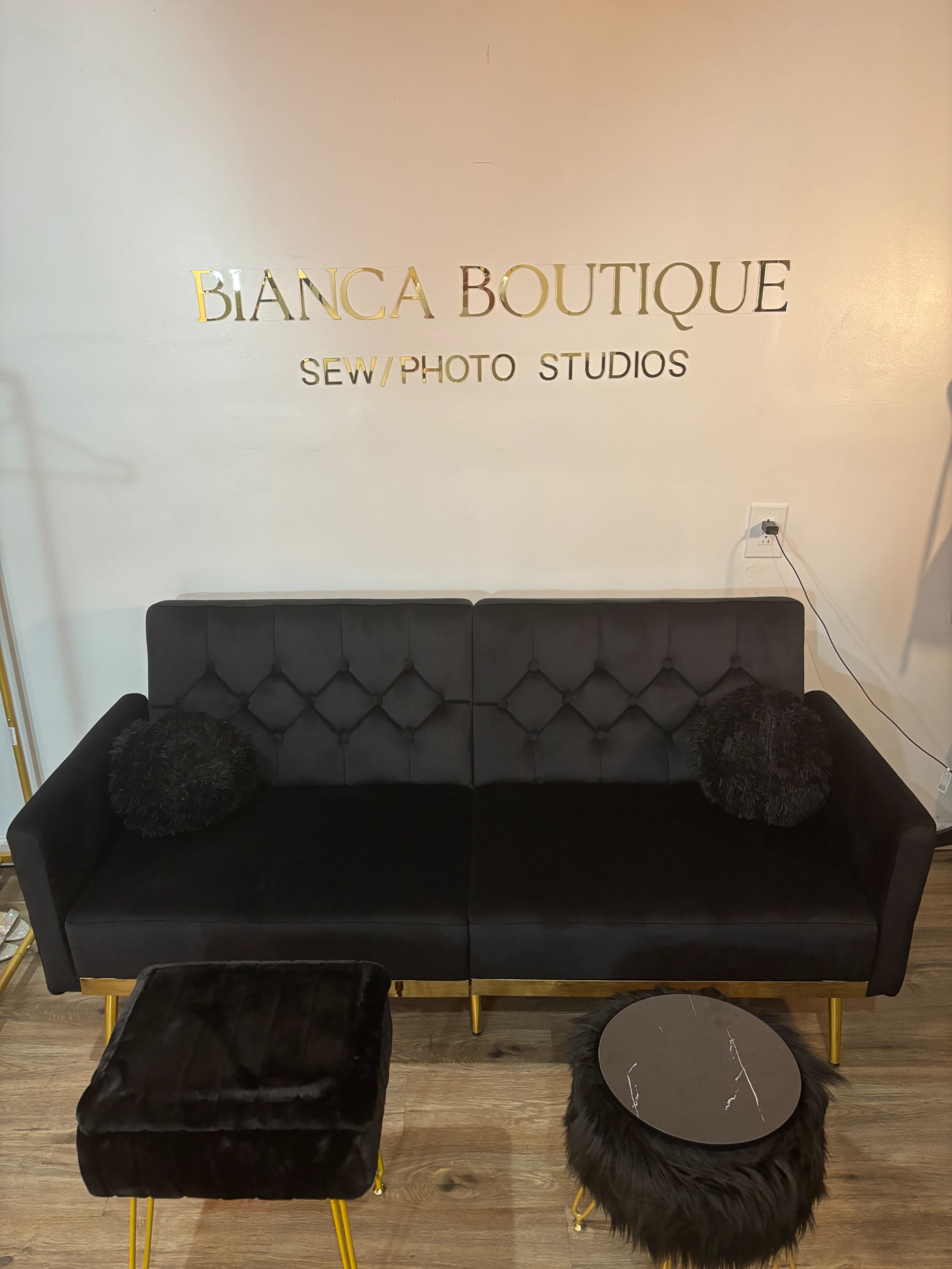 The image shows a black couch with tufted upholstery and decorative cushions, positioned beneath a gold sign that reads "BIANCA BOUTIQUE SEW/PHOTO STUDIOS."