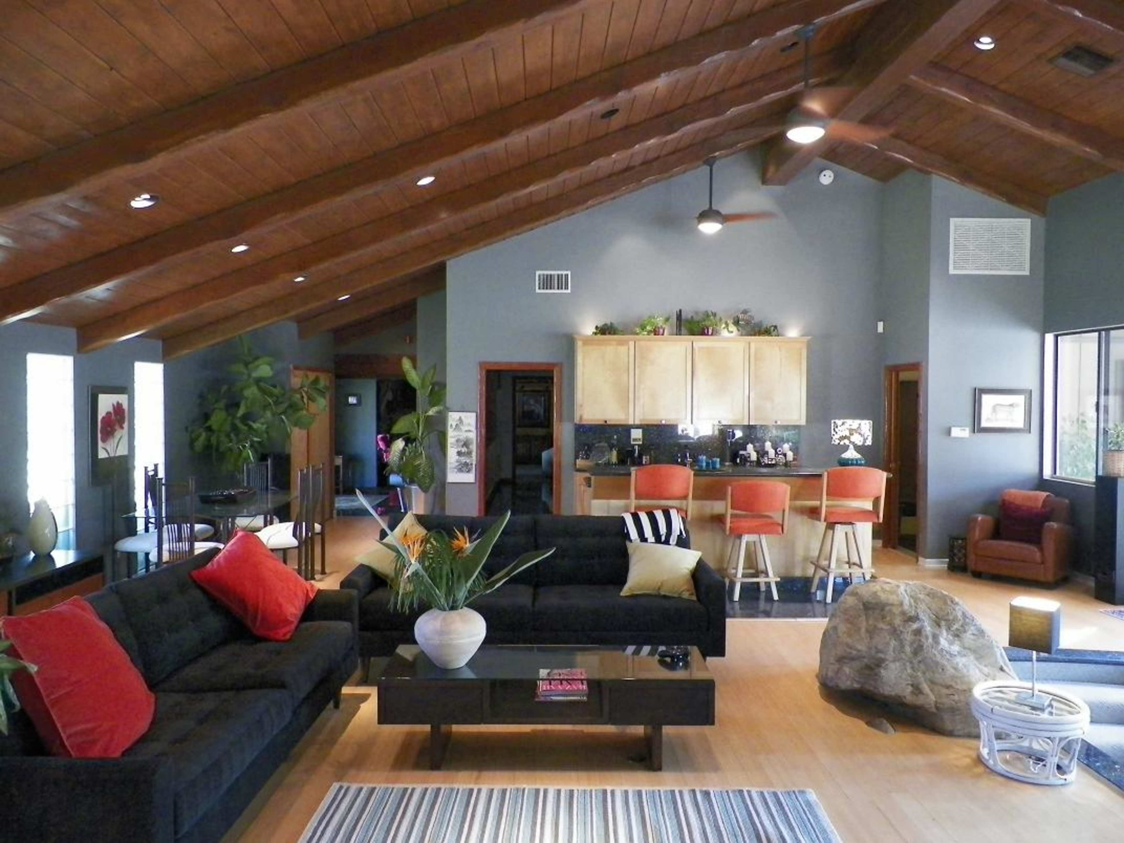 A spacious living area features black sofas with red pillows, a wooden coffee table, and a kitchen bar in the background.