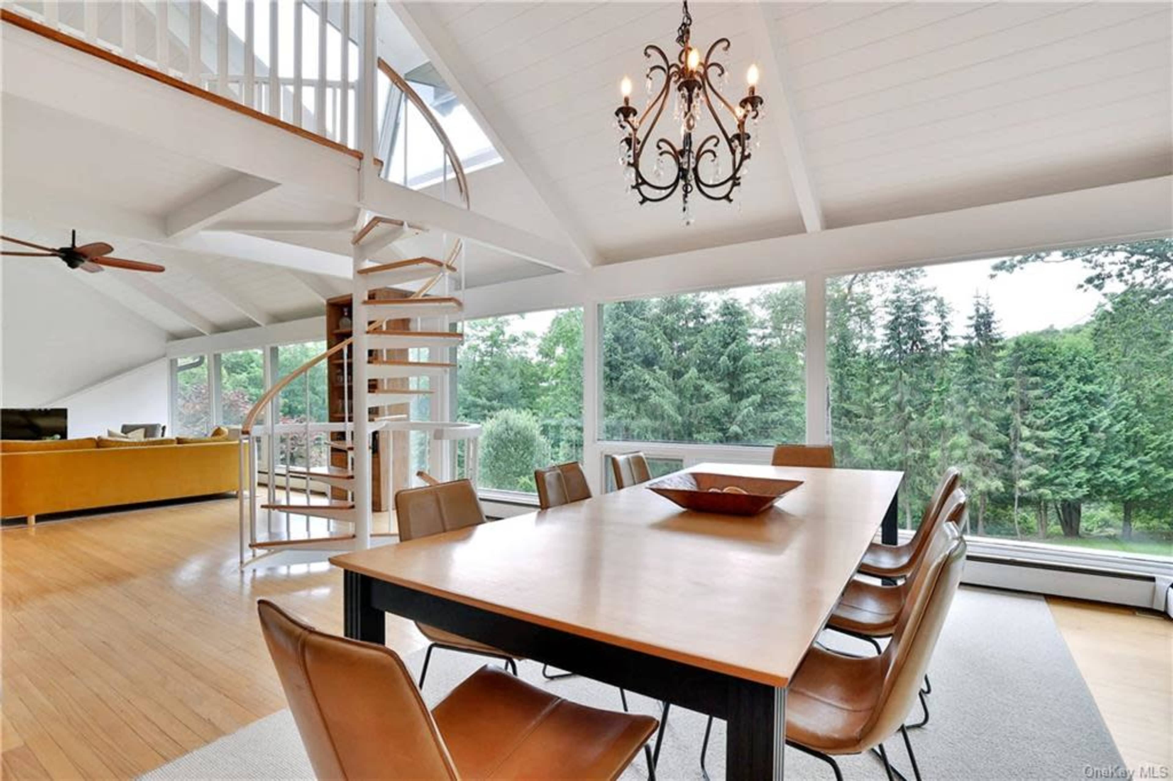 A modern dining area features a long table surrounded by leather chairs, with large windows offering lush outdoor views and a spiral staircase leading to an upper level.
