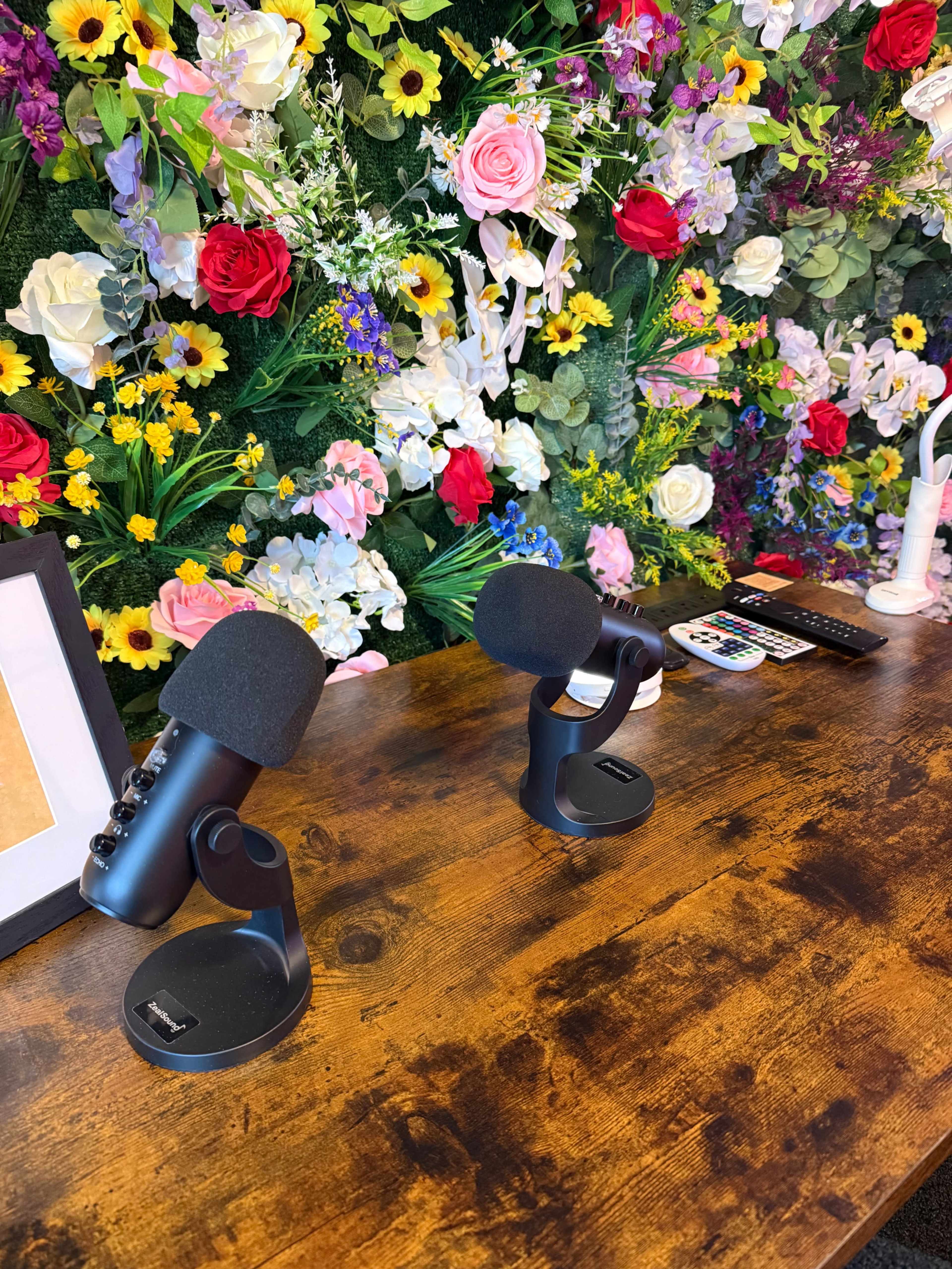 Two black microphones are positioned on a wooden table in front of a colorful flower wall.