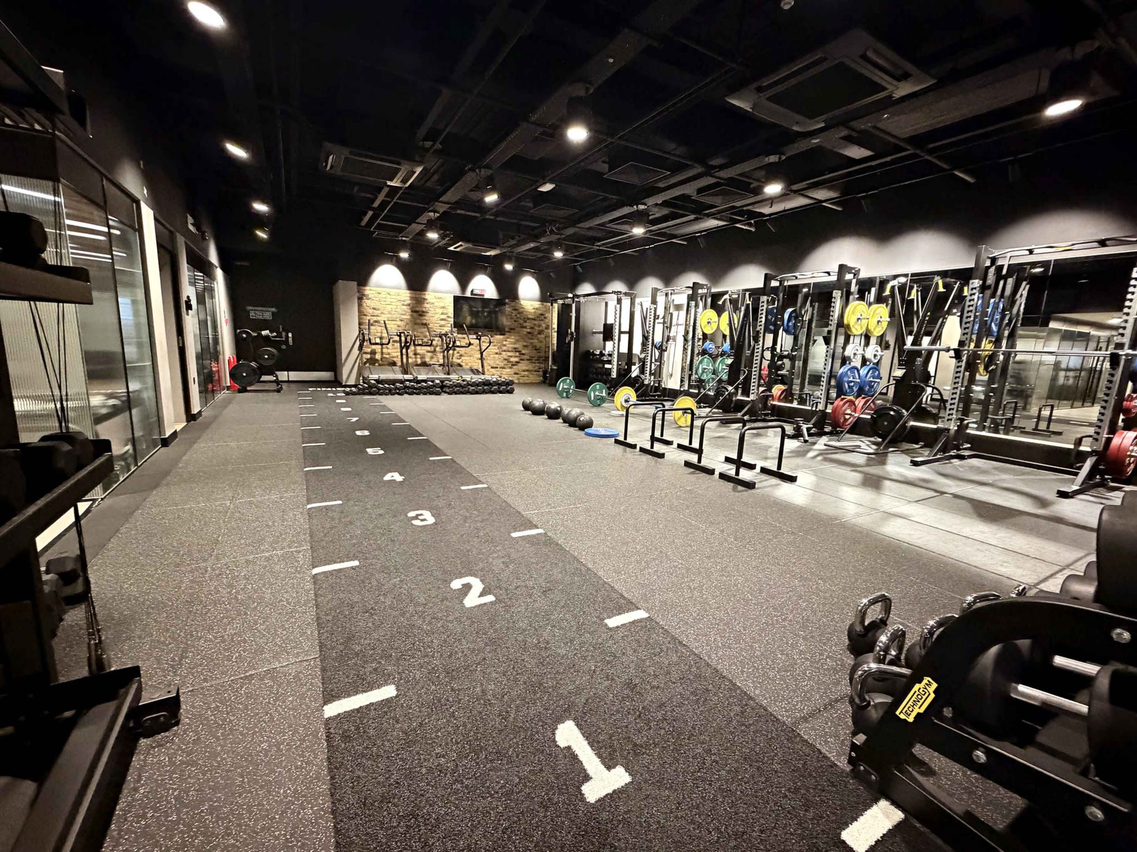 The image shows a modern gym interior with weightlifting equipment, a sprint track marked with numbers, and various exercise tools arranged neatly along the walls.