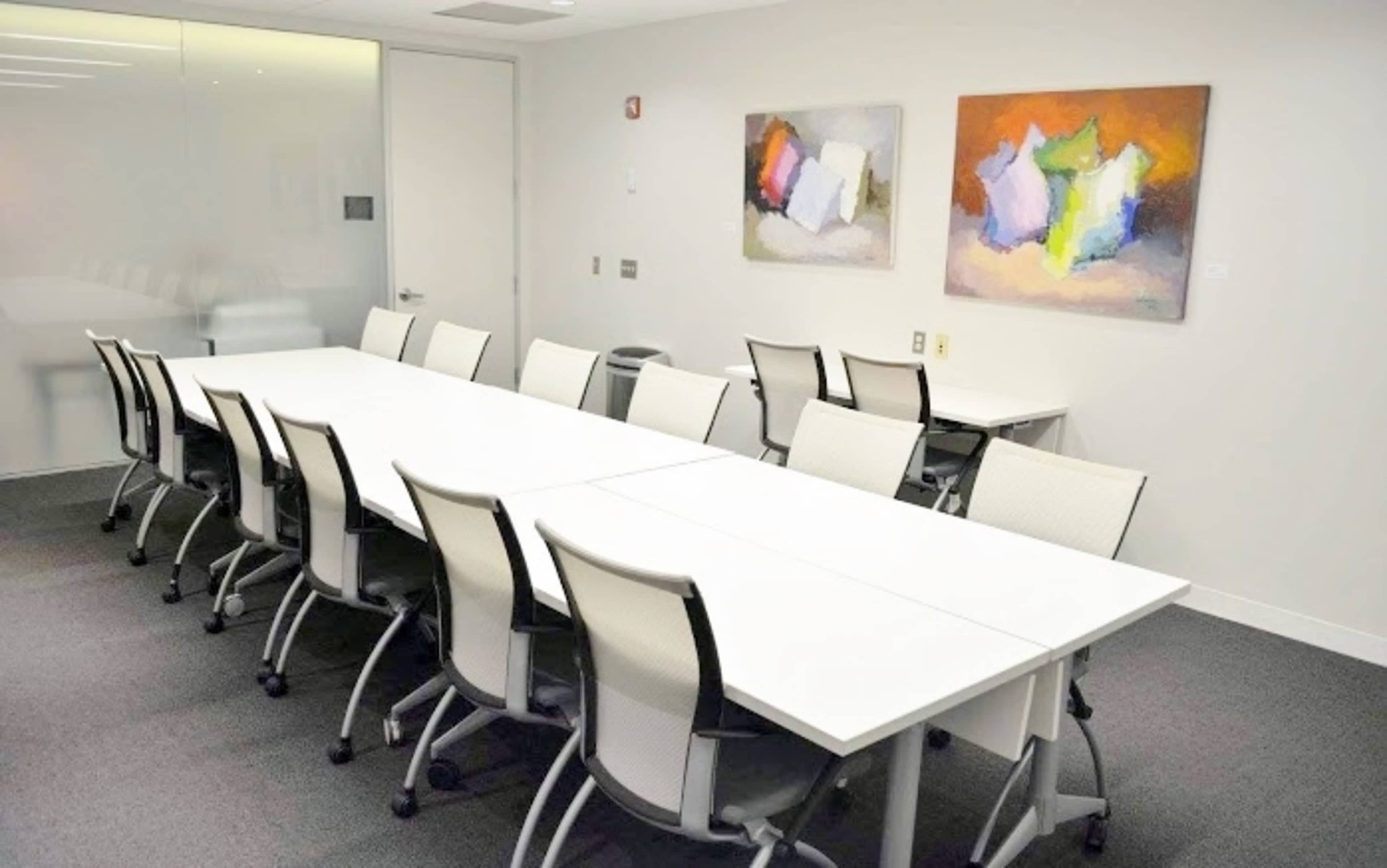 A conference room features a long white table surrounded by several office chairs, with abstract paintings on the walls.