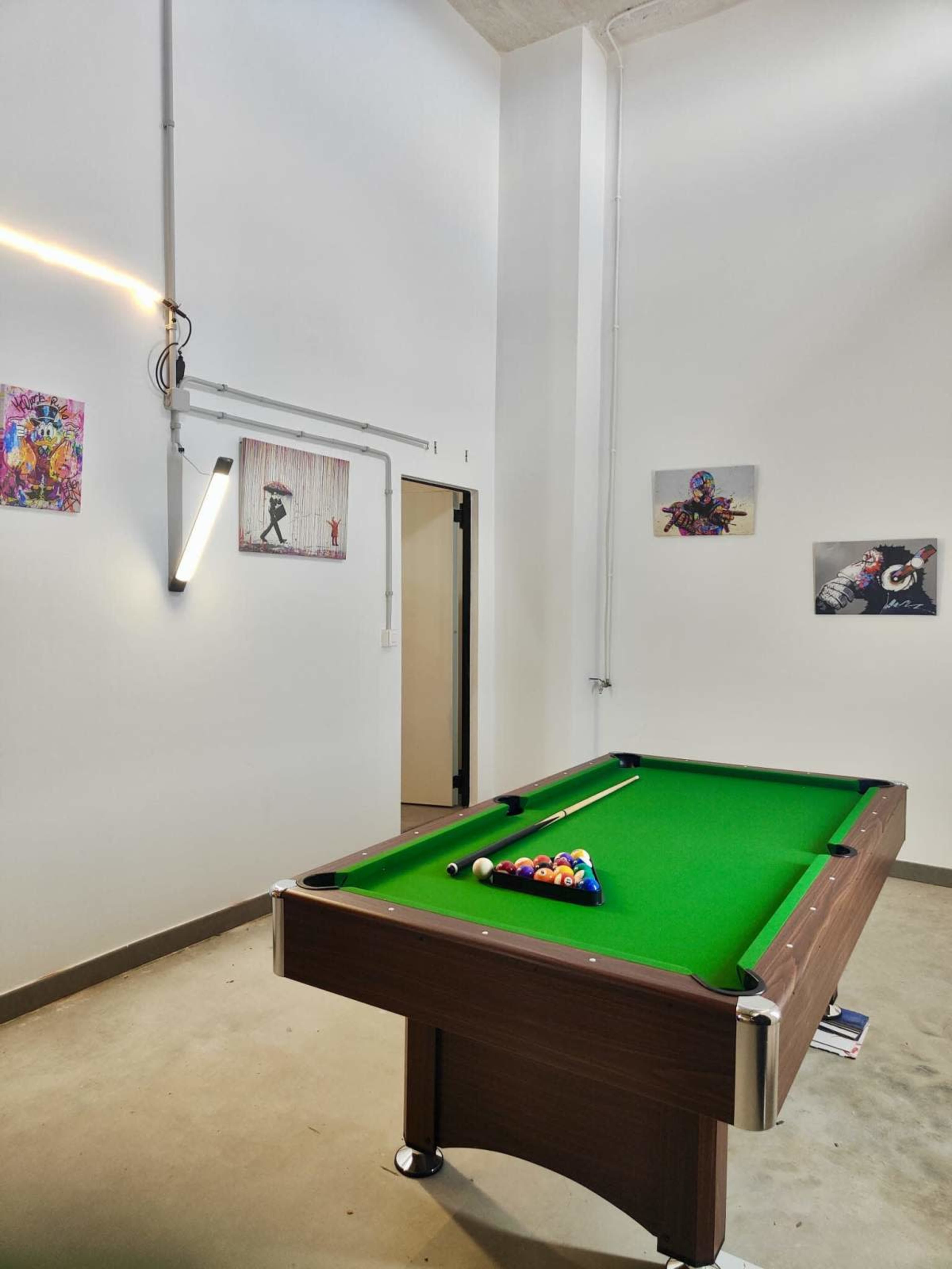 A green pool table is positioned in a room with white walls and several framed artworks hanging, along with exposed wiring and a light fixture.