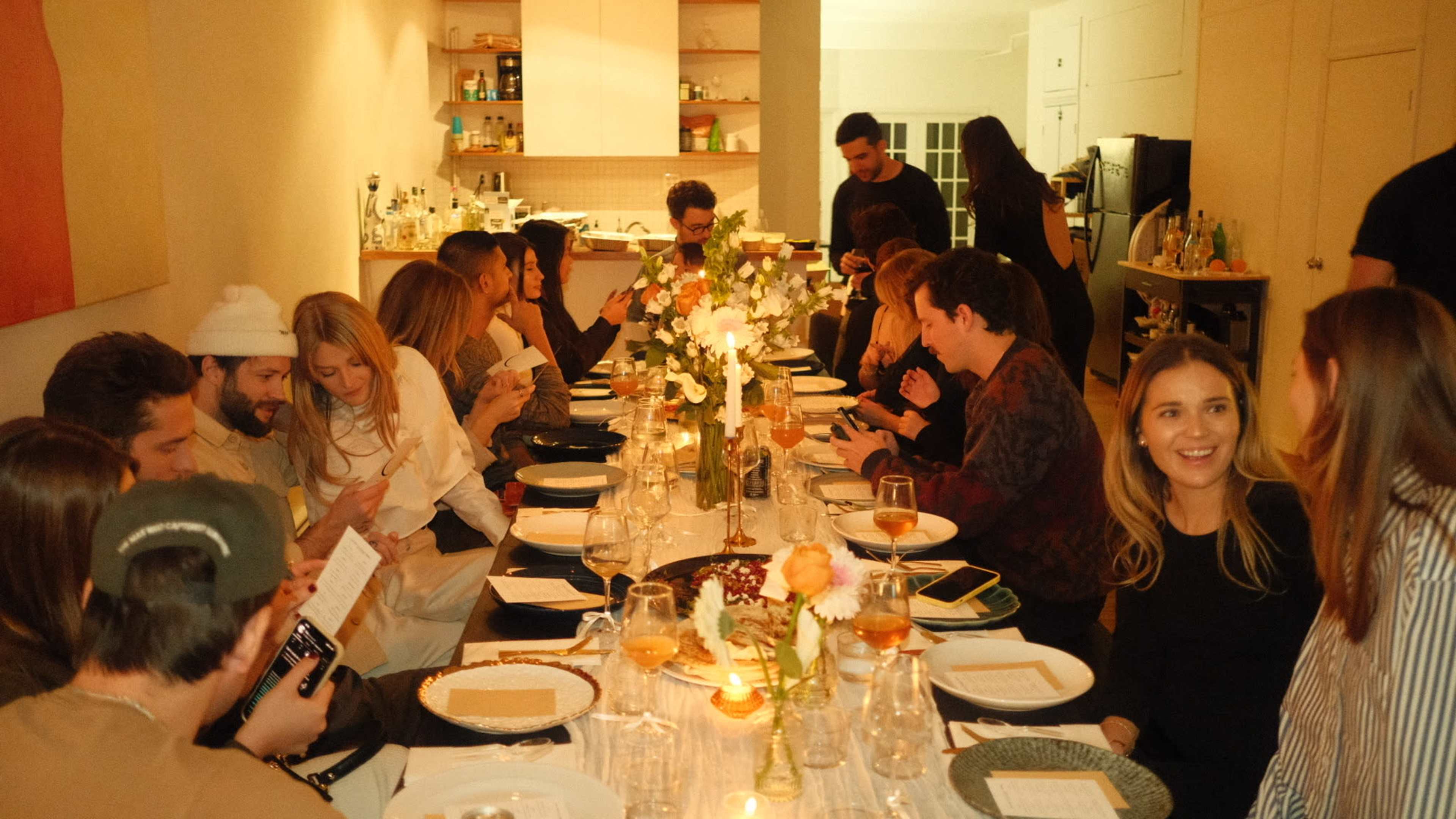 A large group of people sits around a long dining table set with plates and glasses in a warmly lit room.