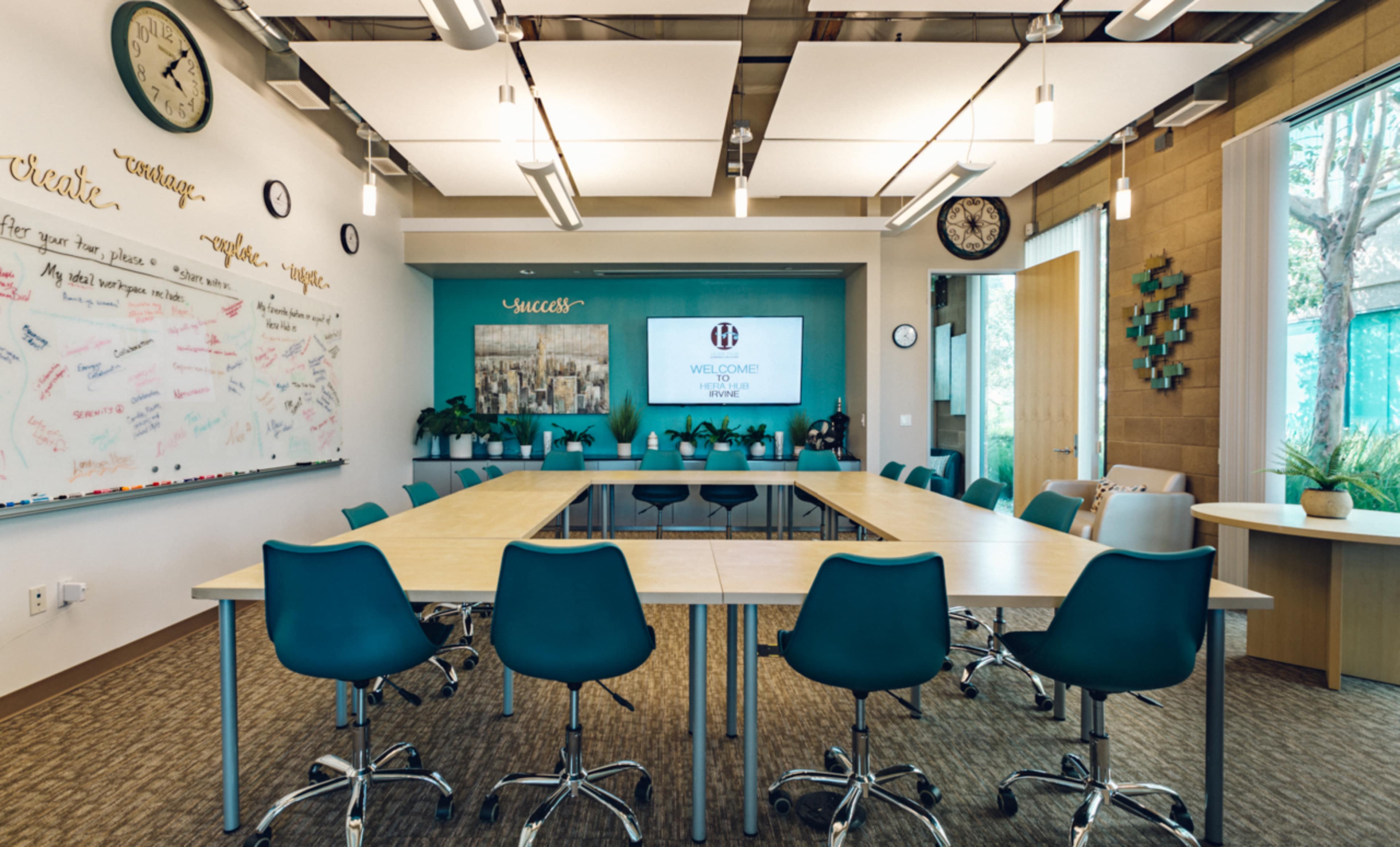 A modern conference room with a long rectangular table surrounded by teal chairs, featuring a large screen, whiteboard covered in writing, and plants along the windows.