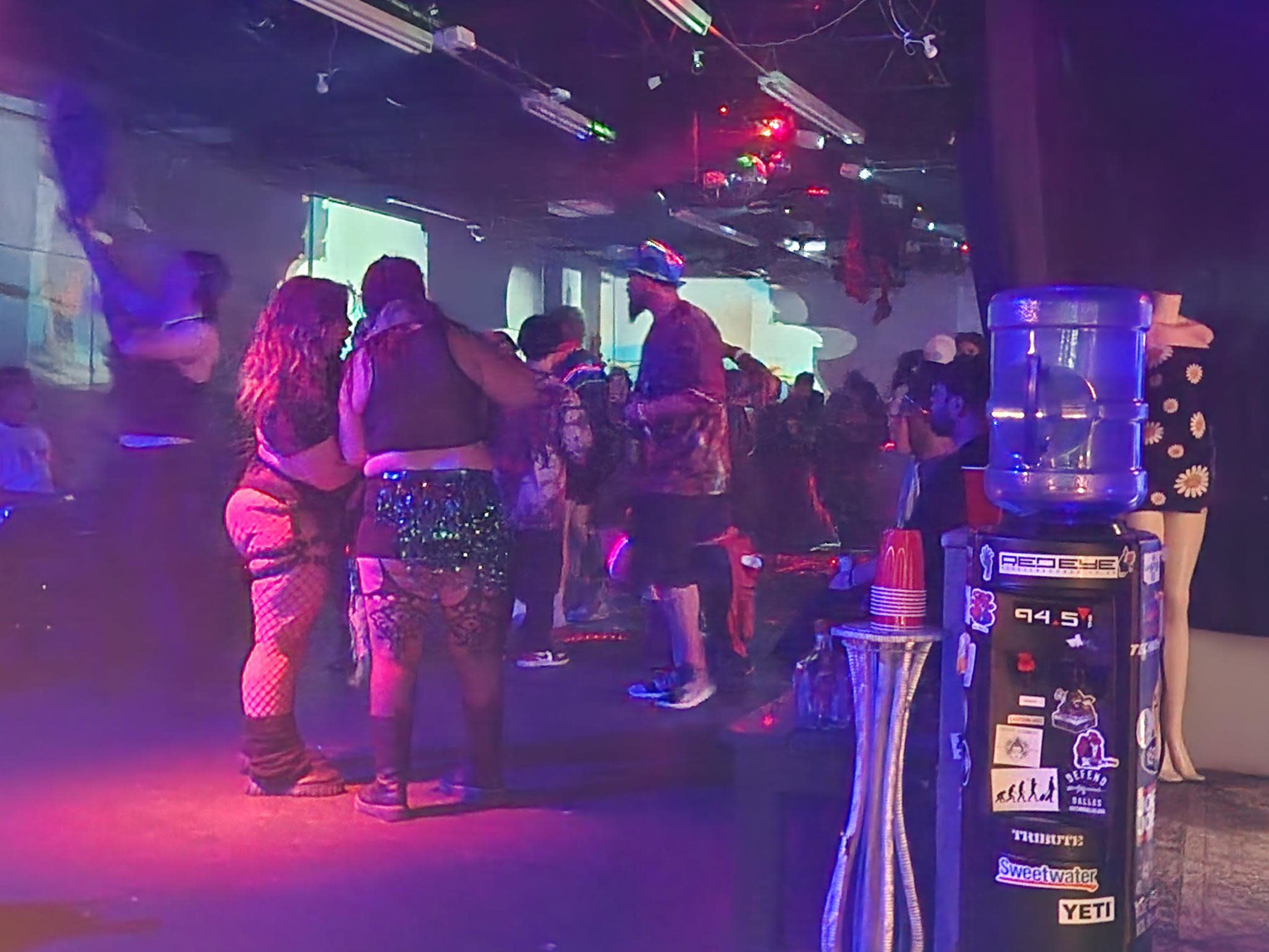 A group of people dance and socialize in a dimly lit venue with colorful lights and projected visuals on the walls.