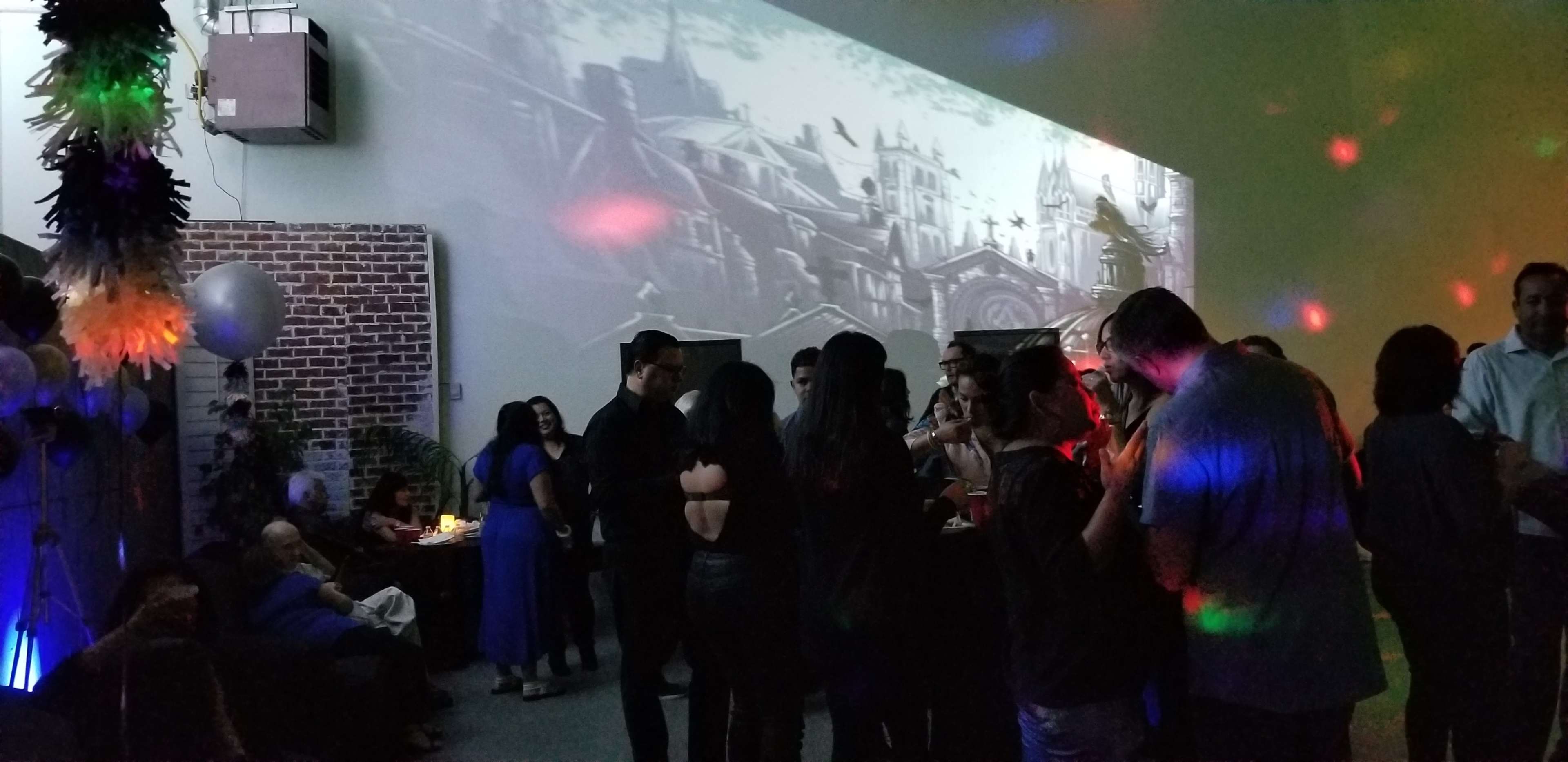 A group of people socialize at a party in a venue with colorful lighting and a large projected mural on the wall.