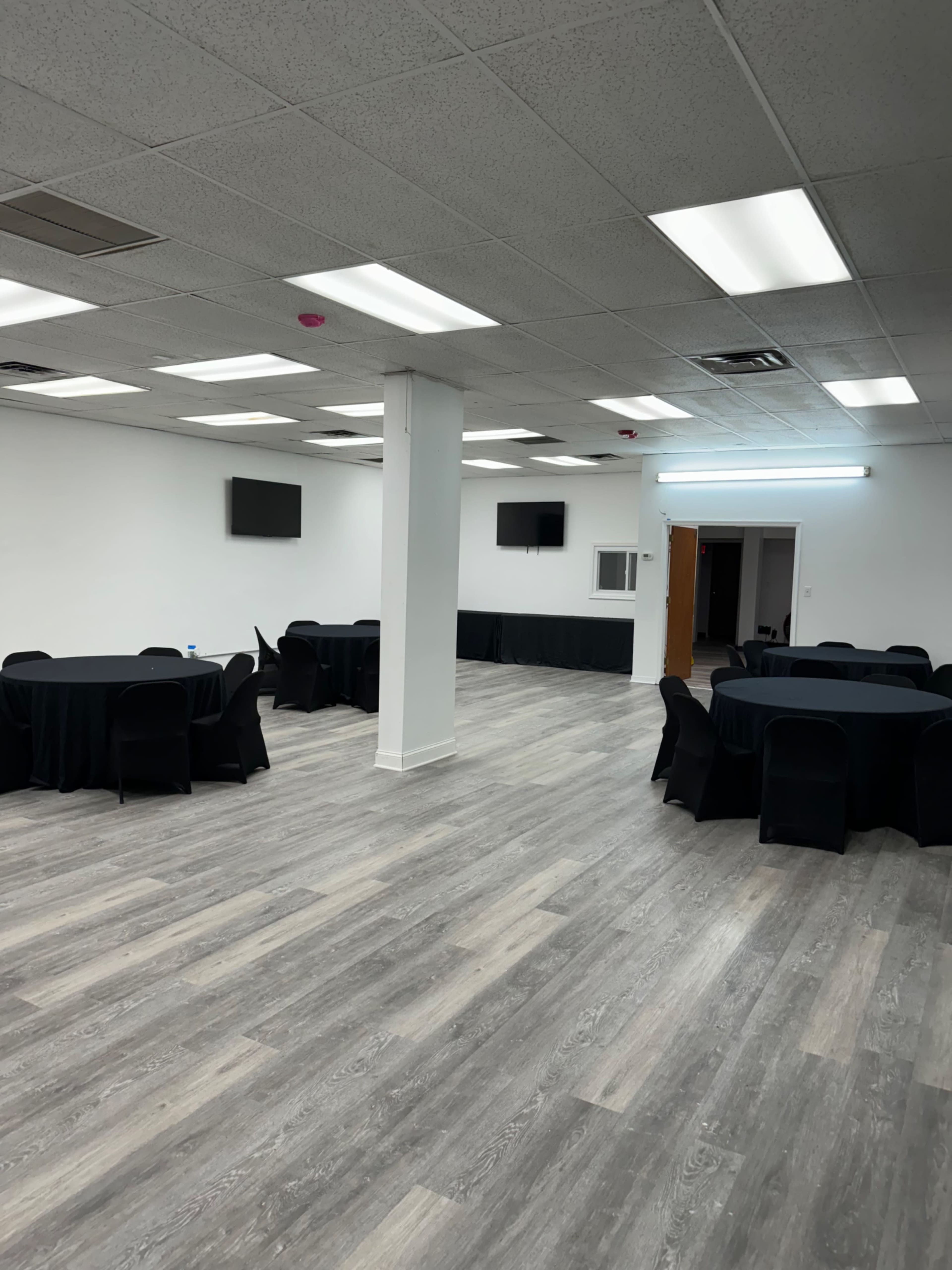 A spacious, empty room with several round tables covered in black tablecloths and TV screens mounted on the walls.
