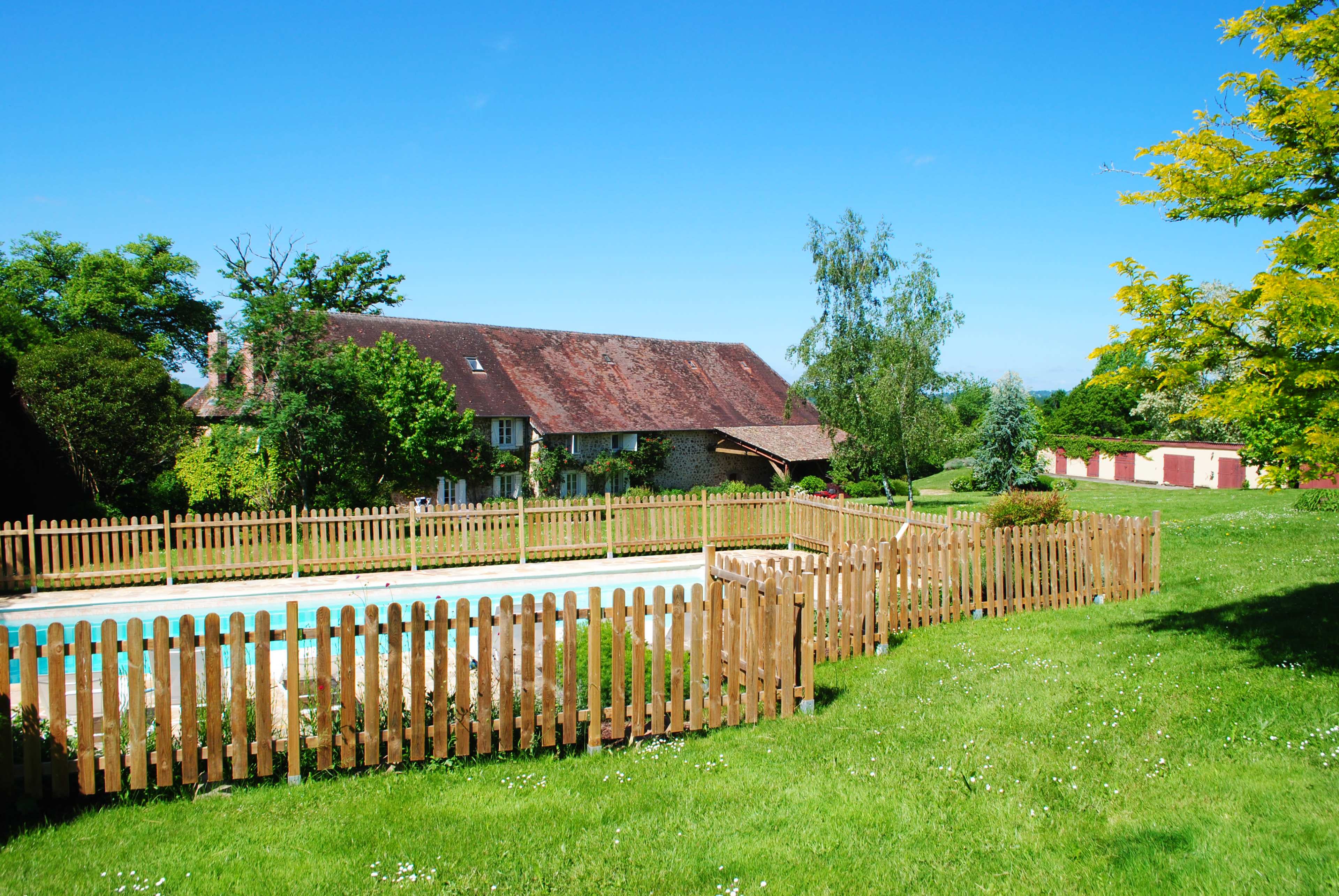 The image shows a grassy area with a fenced swimming pool beside a country house and several trees under a clear blue sky.
