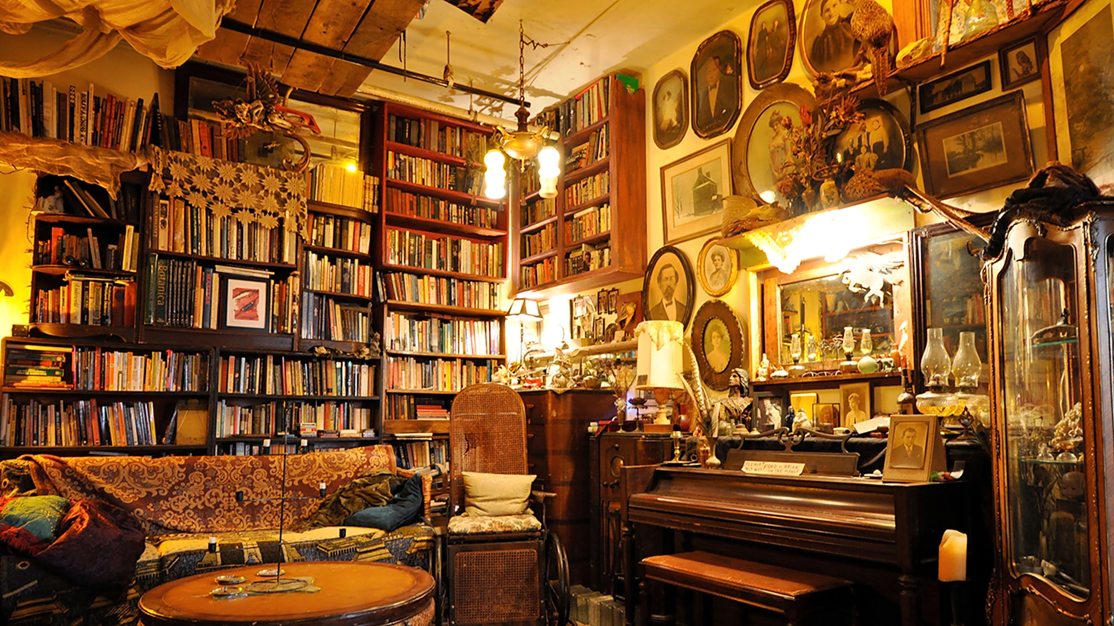 A cozy, vintage-style room filled with bookshelves, antique furniture, and framed artworks, including a piano and various decorative items.