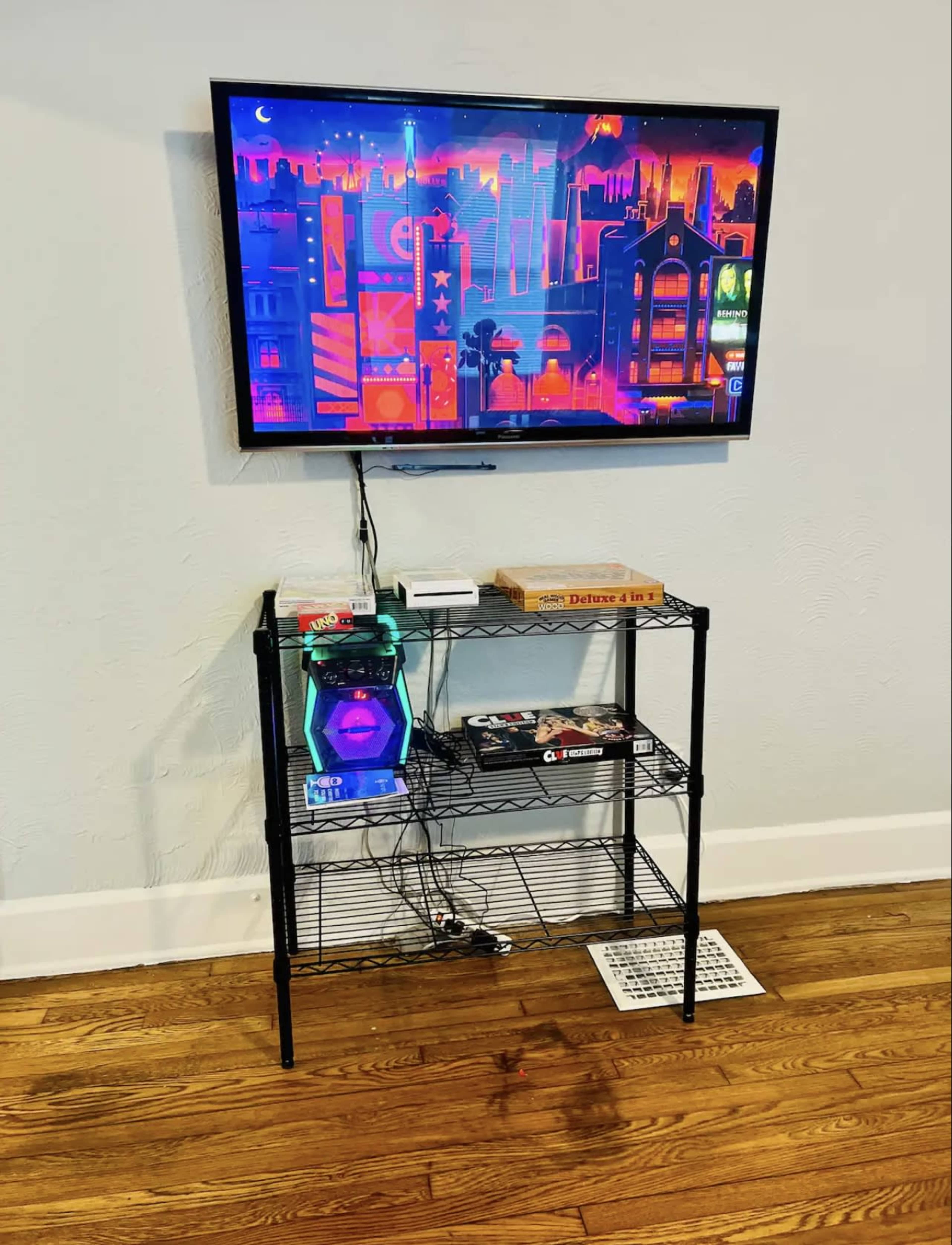 A black wire shelving unit holds a game console and magazines beneath a mounted television displaying a colorful urban skyline.