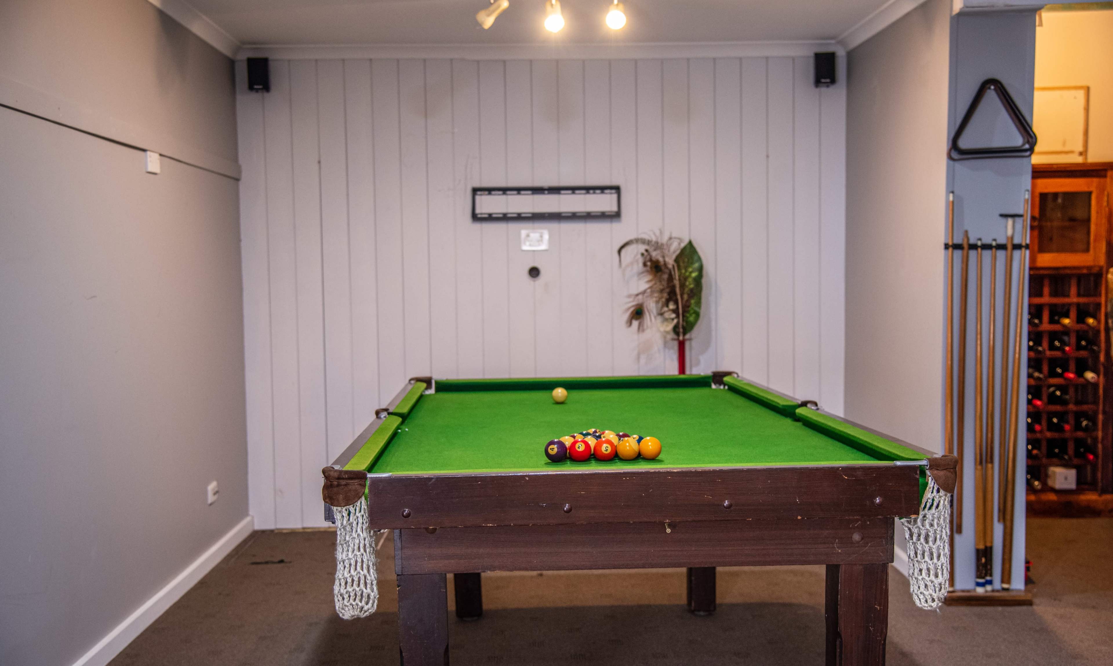 A green billiard table with a set of pool balls arranged on it is positioned in a room featuring pale walls and a small plant in the corner.