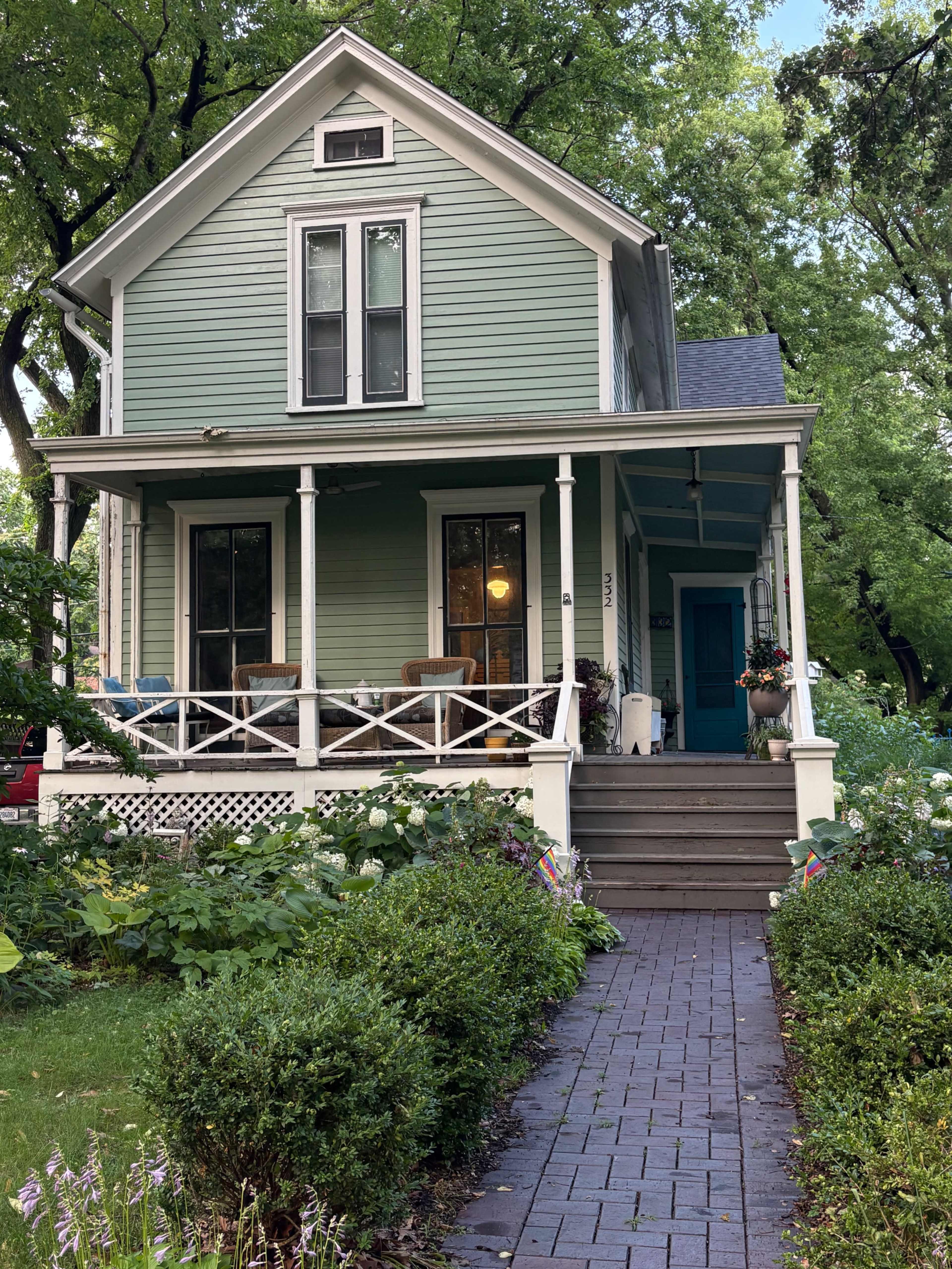 The image features a light green house with a front porch, white railings, and a brick path leading up to it, surrounded by lush greenery and flowers.
