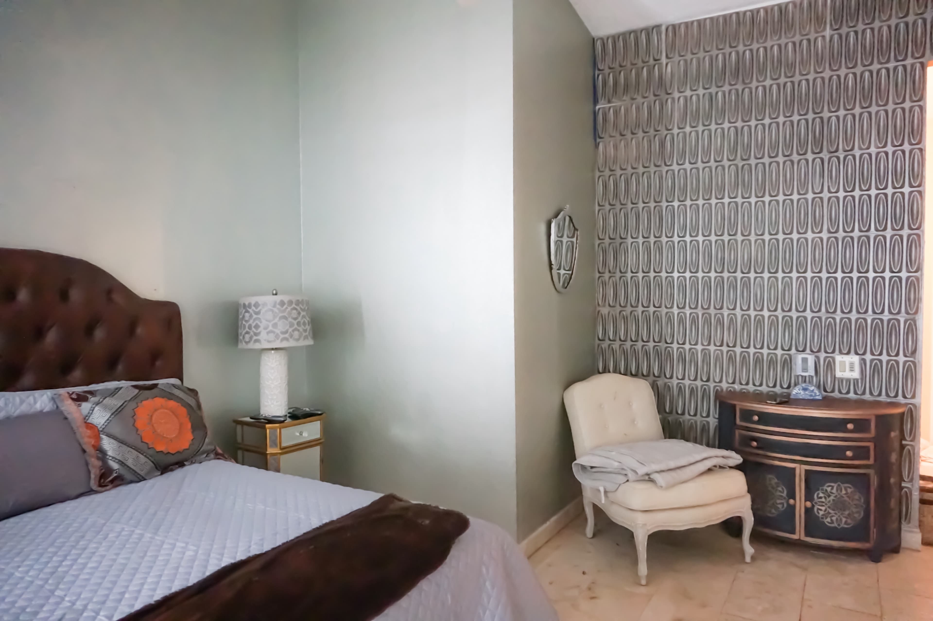 A bedroom with a bed featuring a tufted headboard, a patterned armchair, and a small decorative table against a patterned wall.