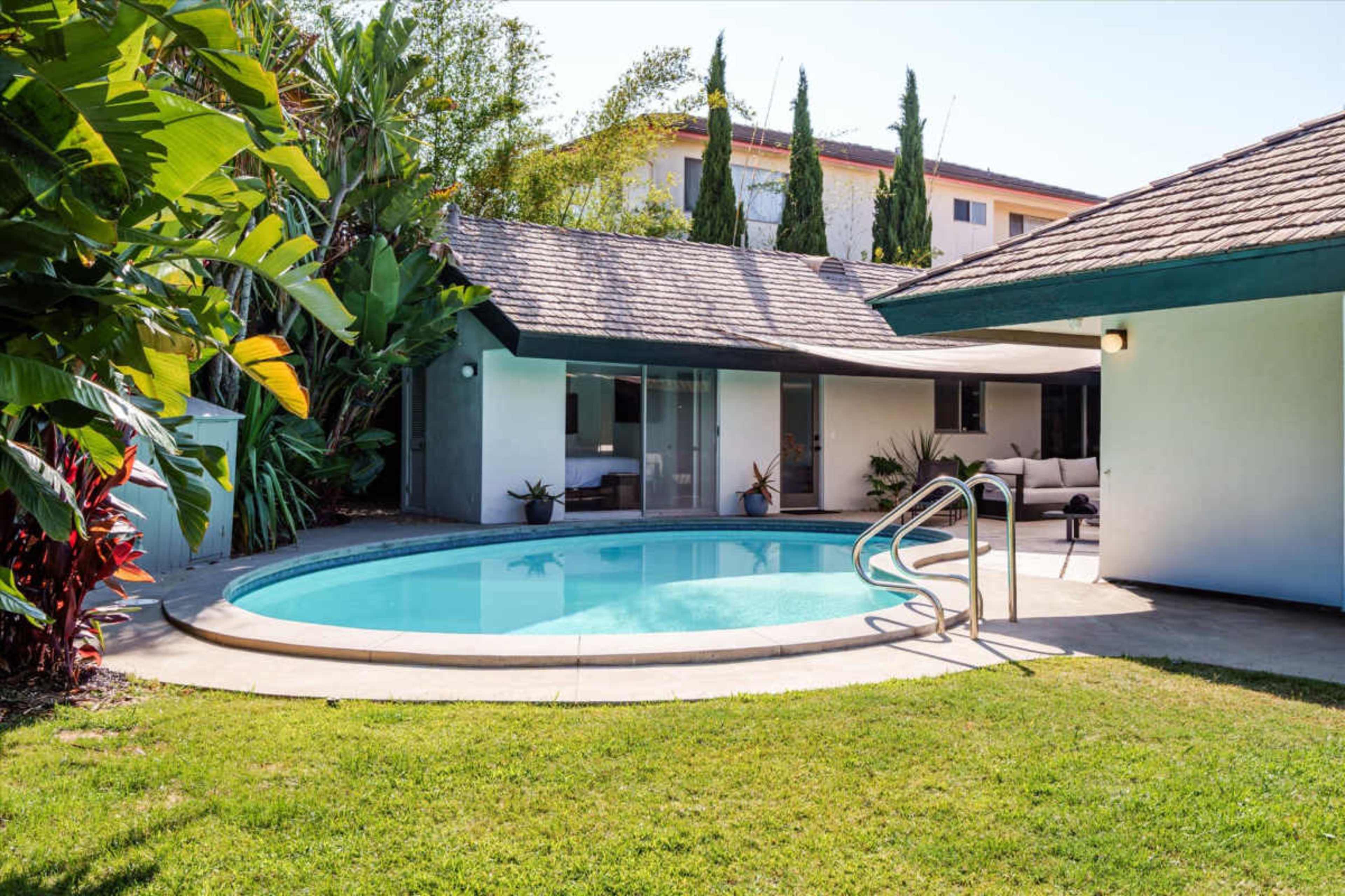 The image shows a landscaped backyard with a circular swimming pool surrounded by grass and tropical plants, next to a modern home.