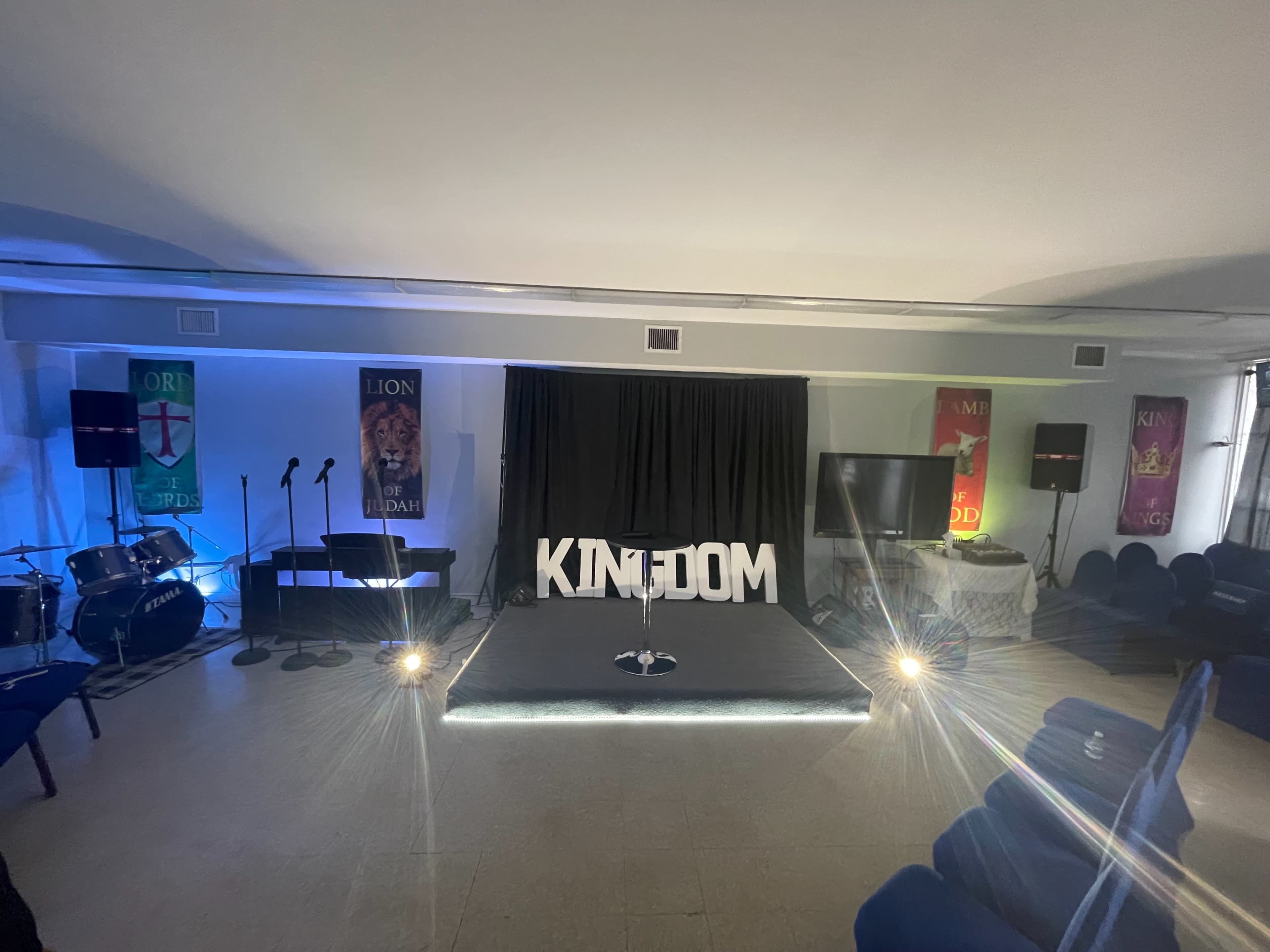 The image depicts a stage setup for a event, featuring the word "KINGDOM" prominently displayed, with banners and musical instruments arranged in a brightly lit room.