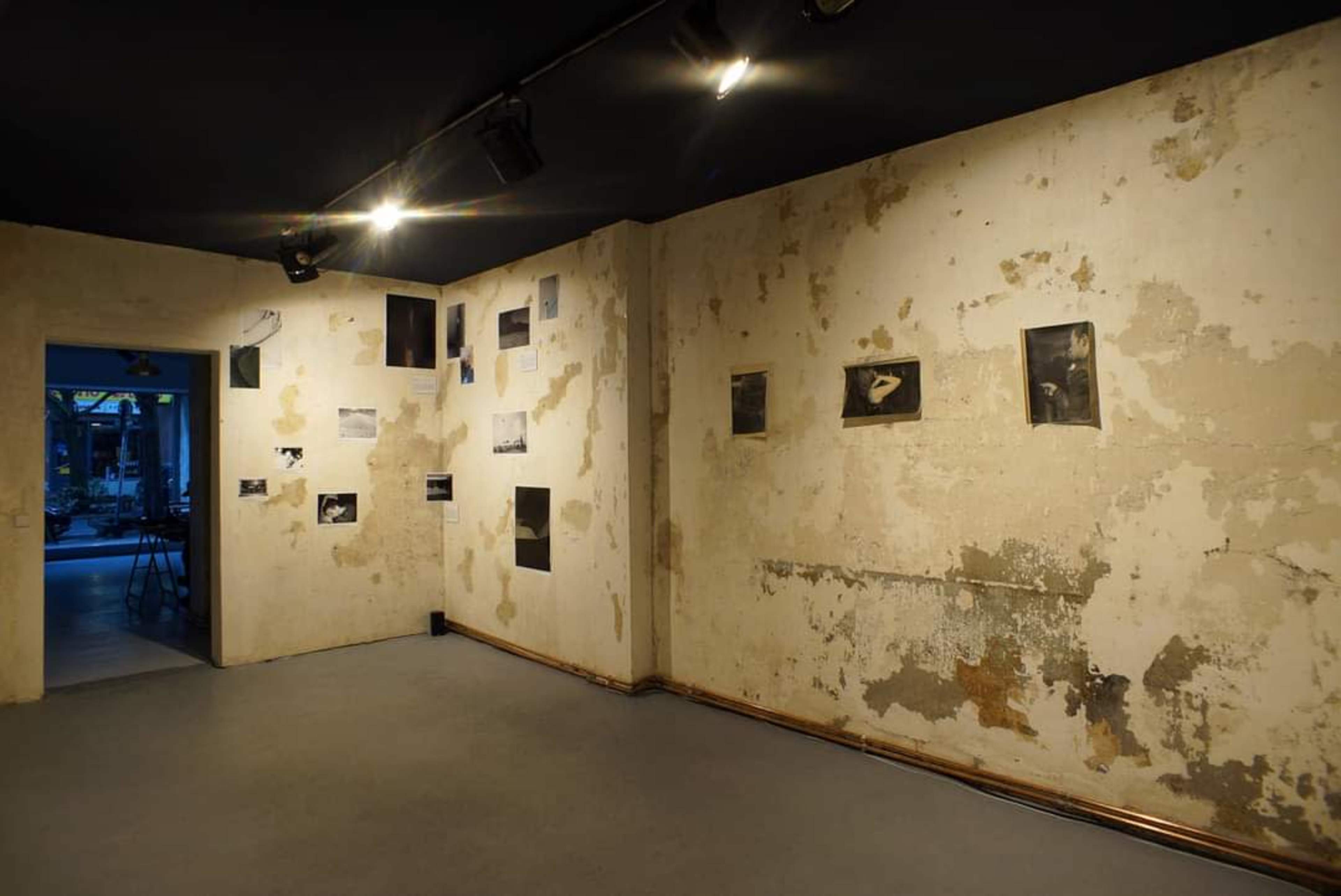The image shows a dimly lit gallery with peeling walls and black-and-white photographs mounted on the walls.