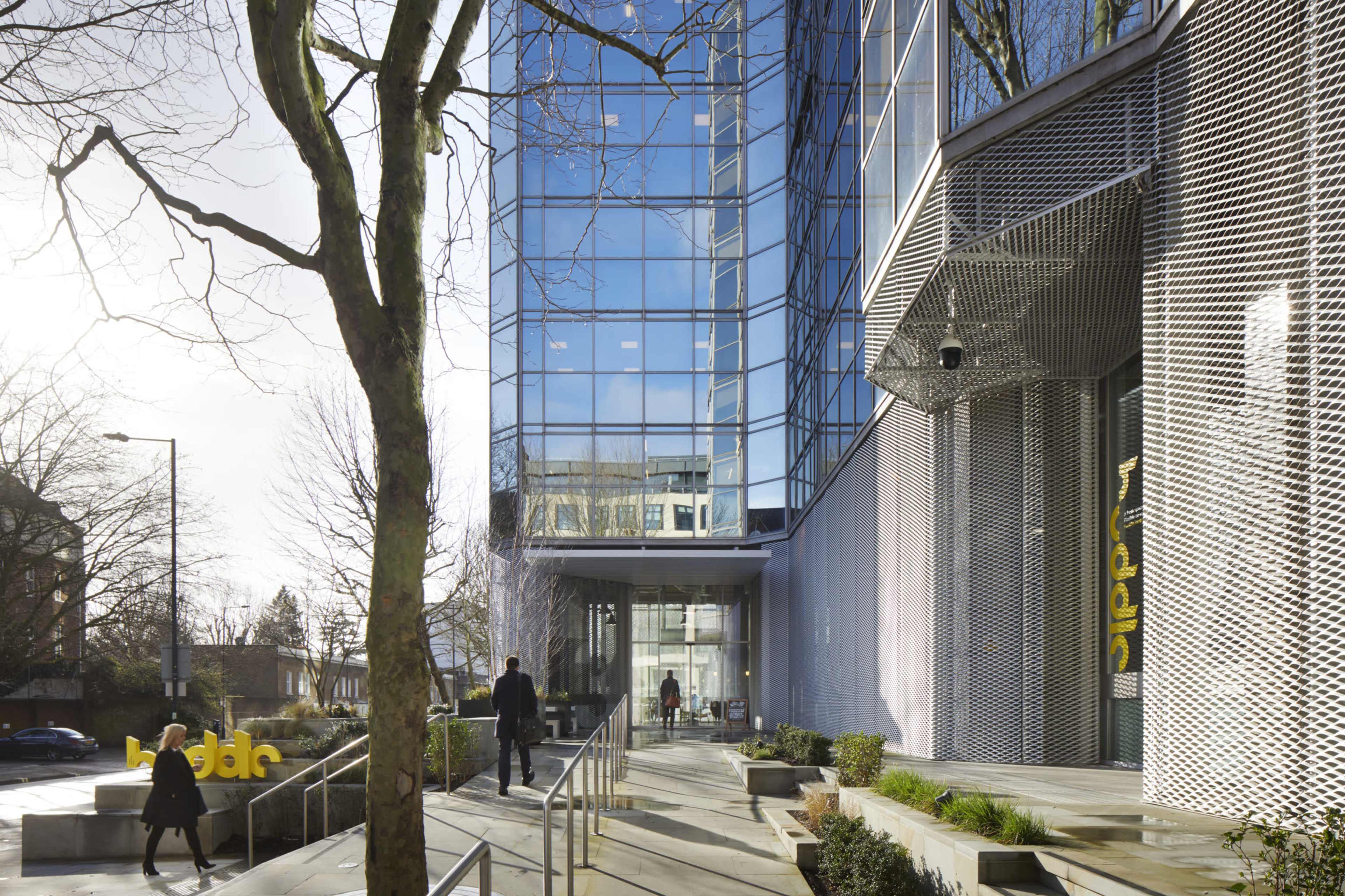 A modern glass building with a structured entrance and people walking along a pathway lined with trees and greenery.