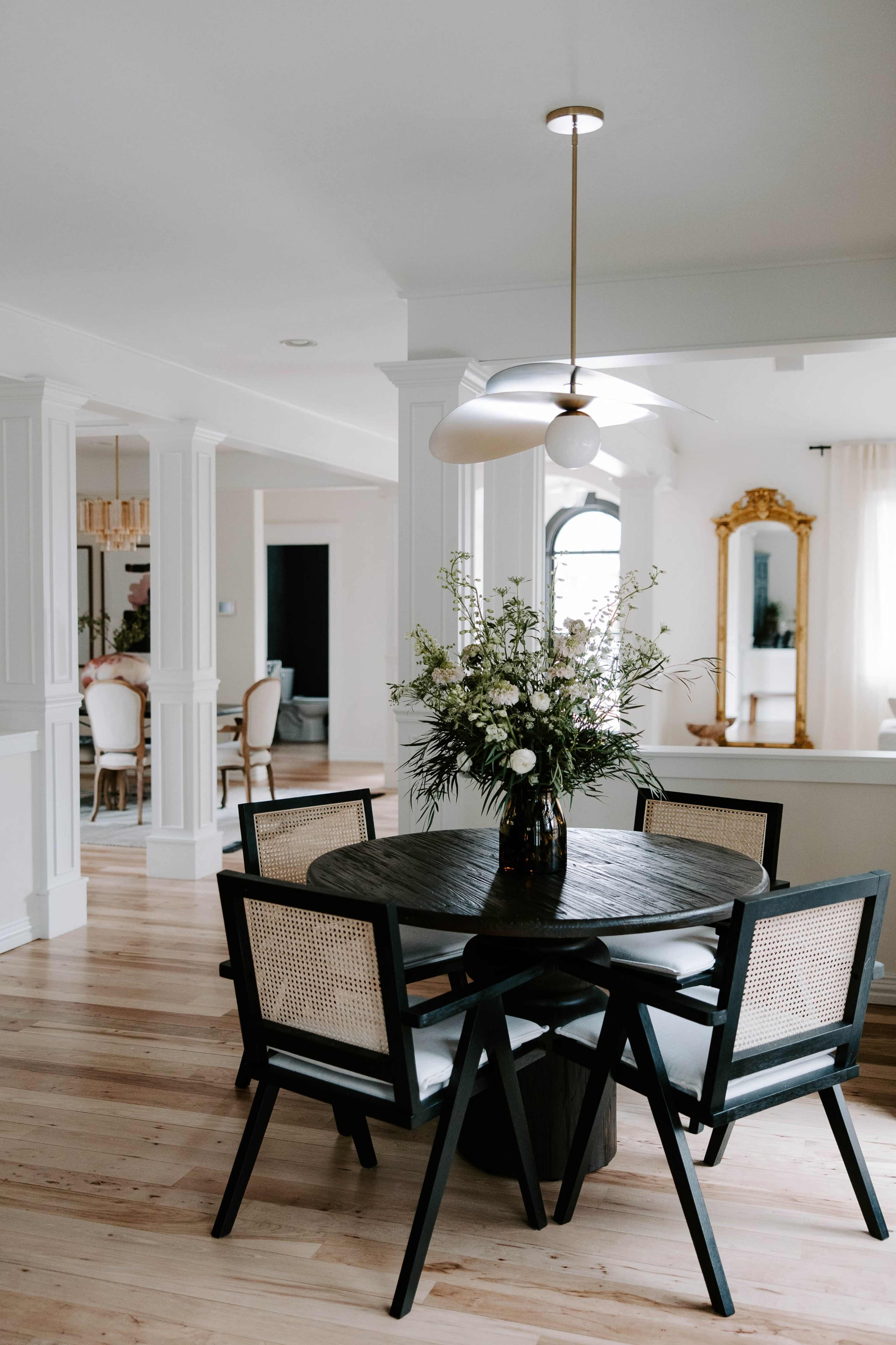 The image shows a modern dining area with a round black table, four black chairs with light cushions, and a vase of flowers, illuminated by a stylish ceiling light.