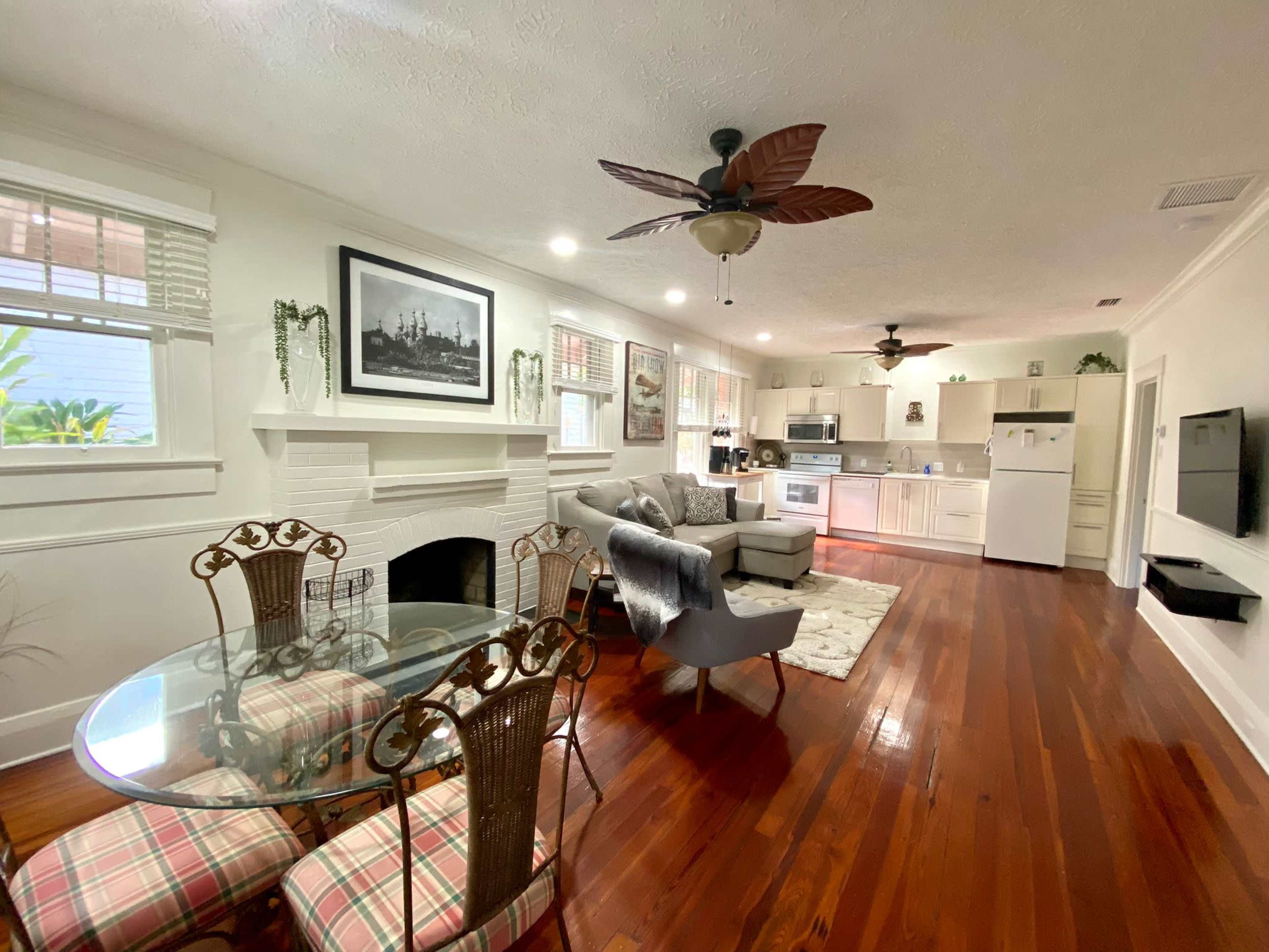 The image shows a well-lit living area featuring a dining table with four chairs, a sofa, and a kitchenette with modern appliances.
