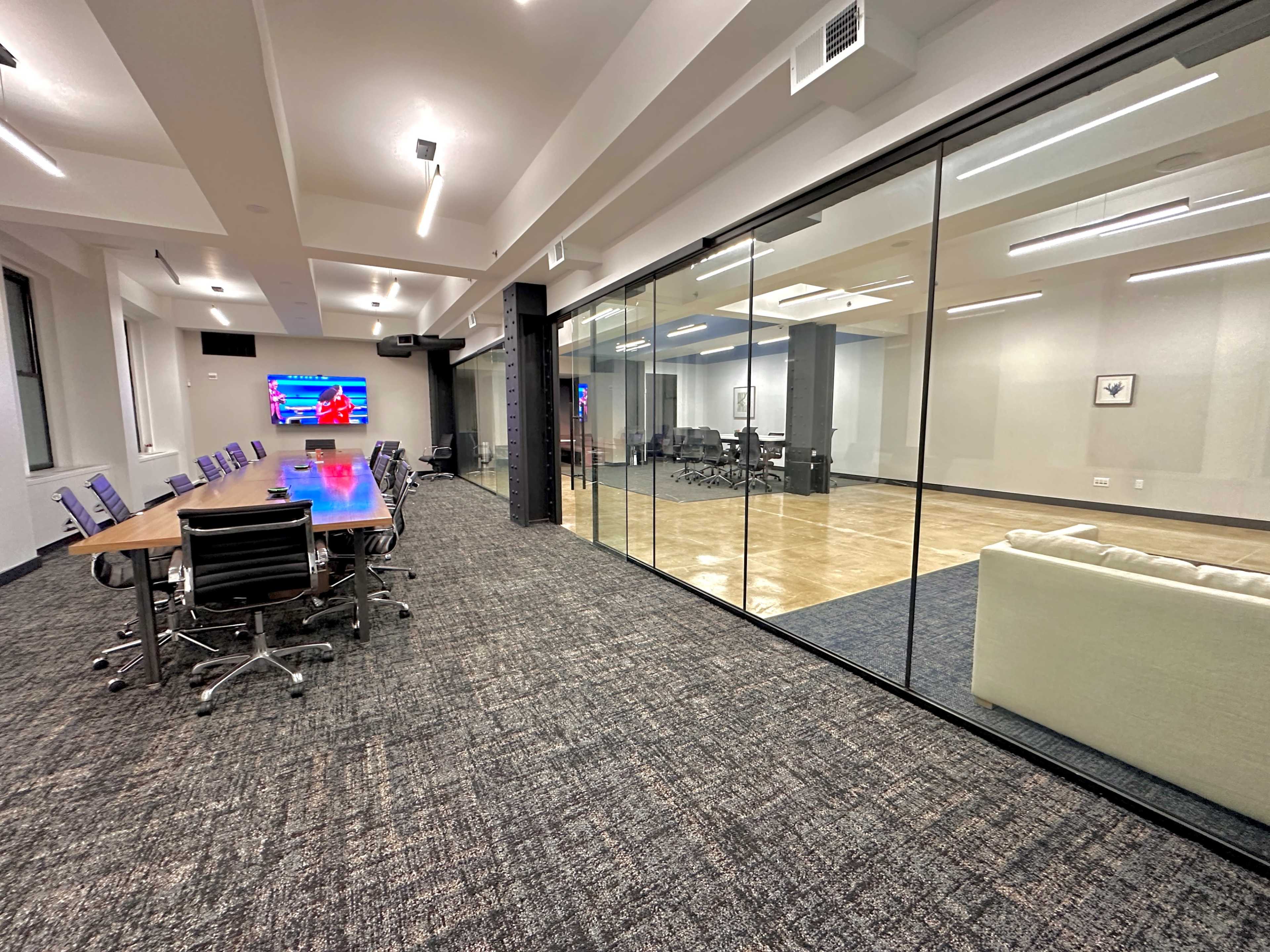 A modern office space features a conference room with a large table and chairs, adjacent to a glass-walled room equipped with additional seating and a presentation screen.