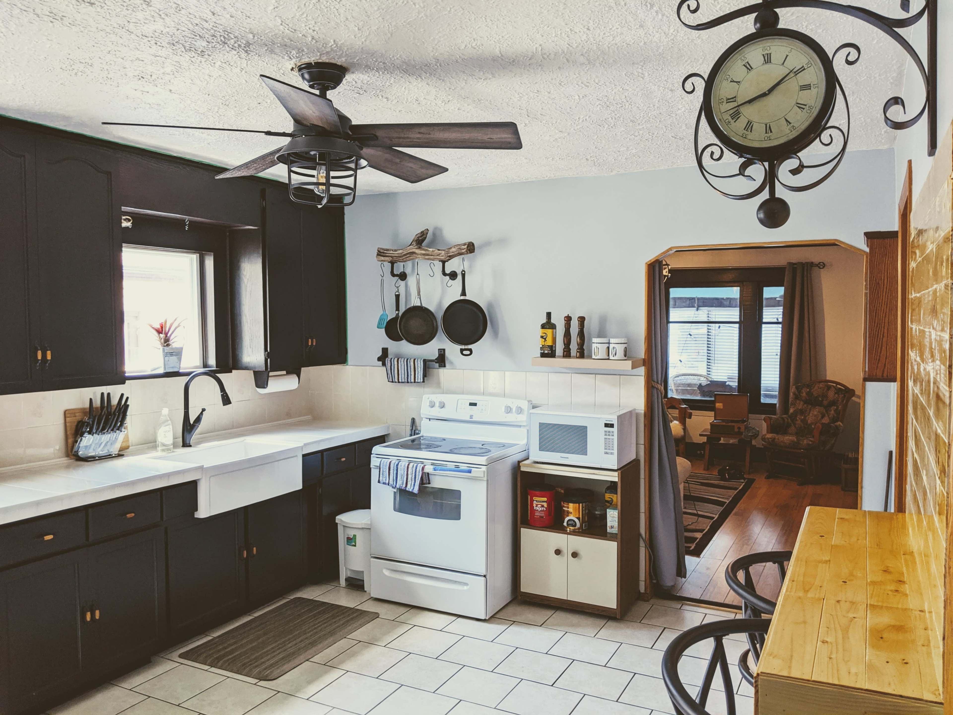 A kitchen with dark cabinets, a white stove, and a countertop equipped with utensils and a microwave, while a clock hangs above and a doorway leads to another room.