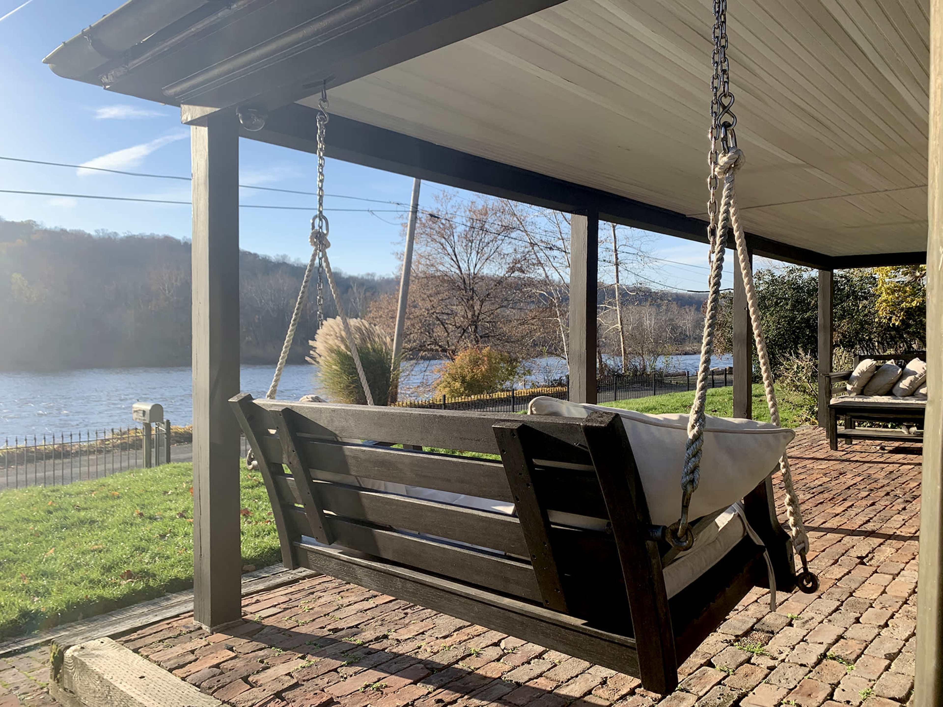 A wooden swing hangs beneath a covered porch overlooking a river with trees in the background.