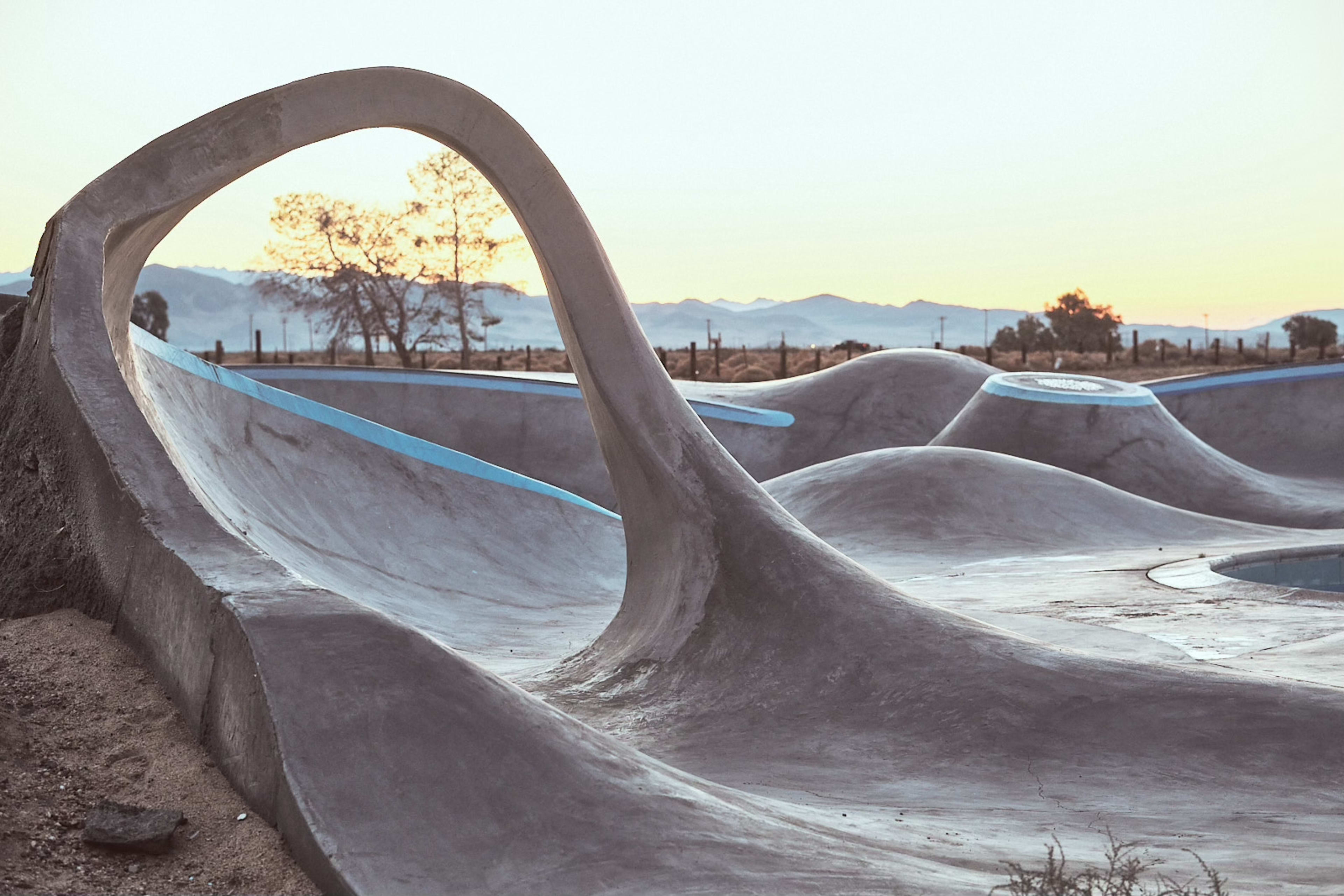 A modern skate park with smooth, flowing concrete contours and a backdrop of distant mountains under a soft sky at sunset.