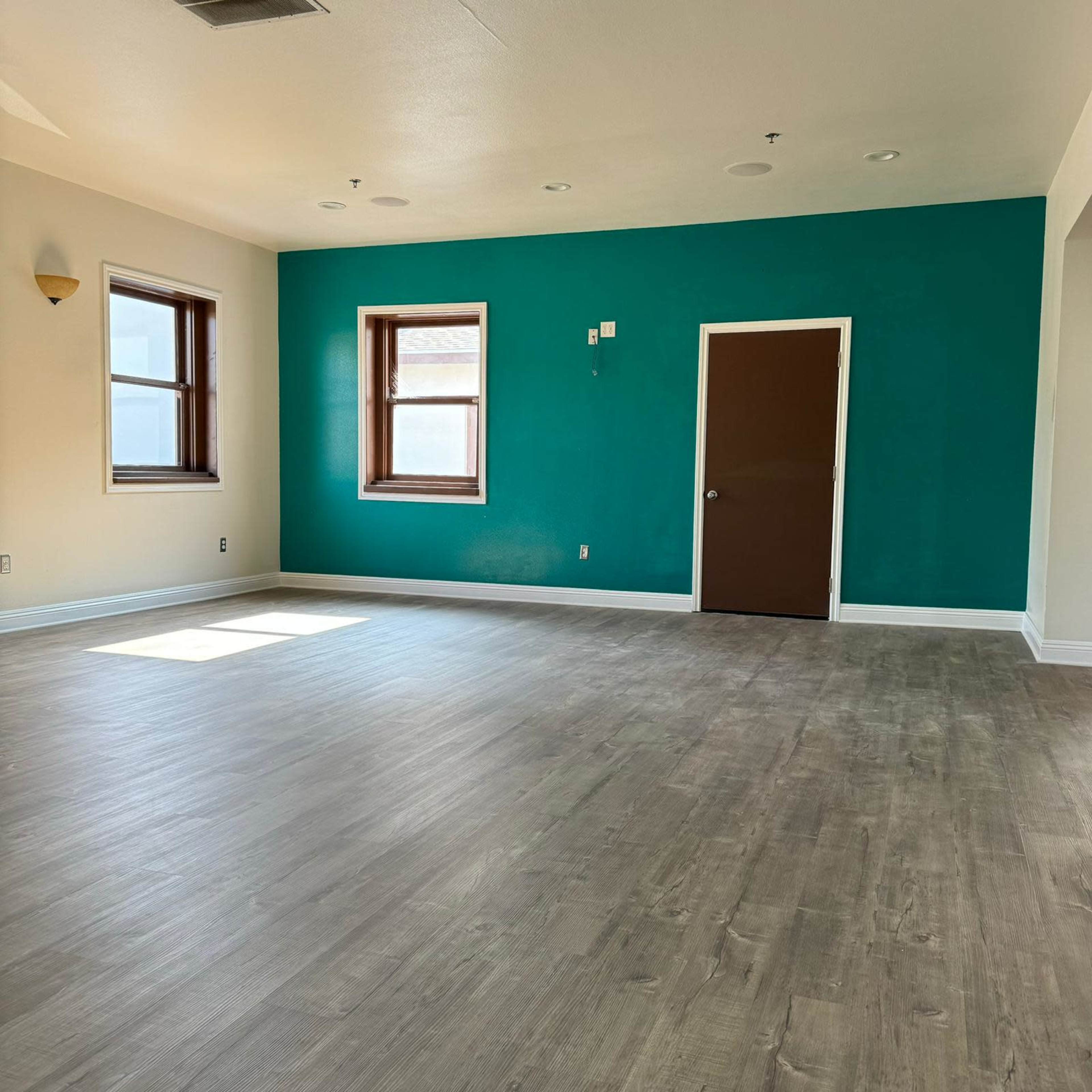 The room features a teal accent wall, two windows, a brown door, and a light-colored vinyl floor.