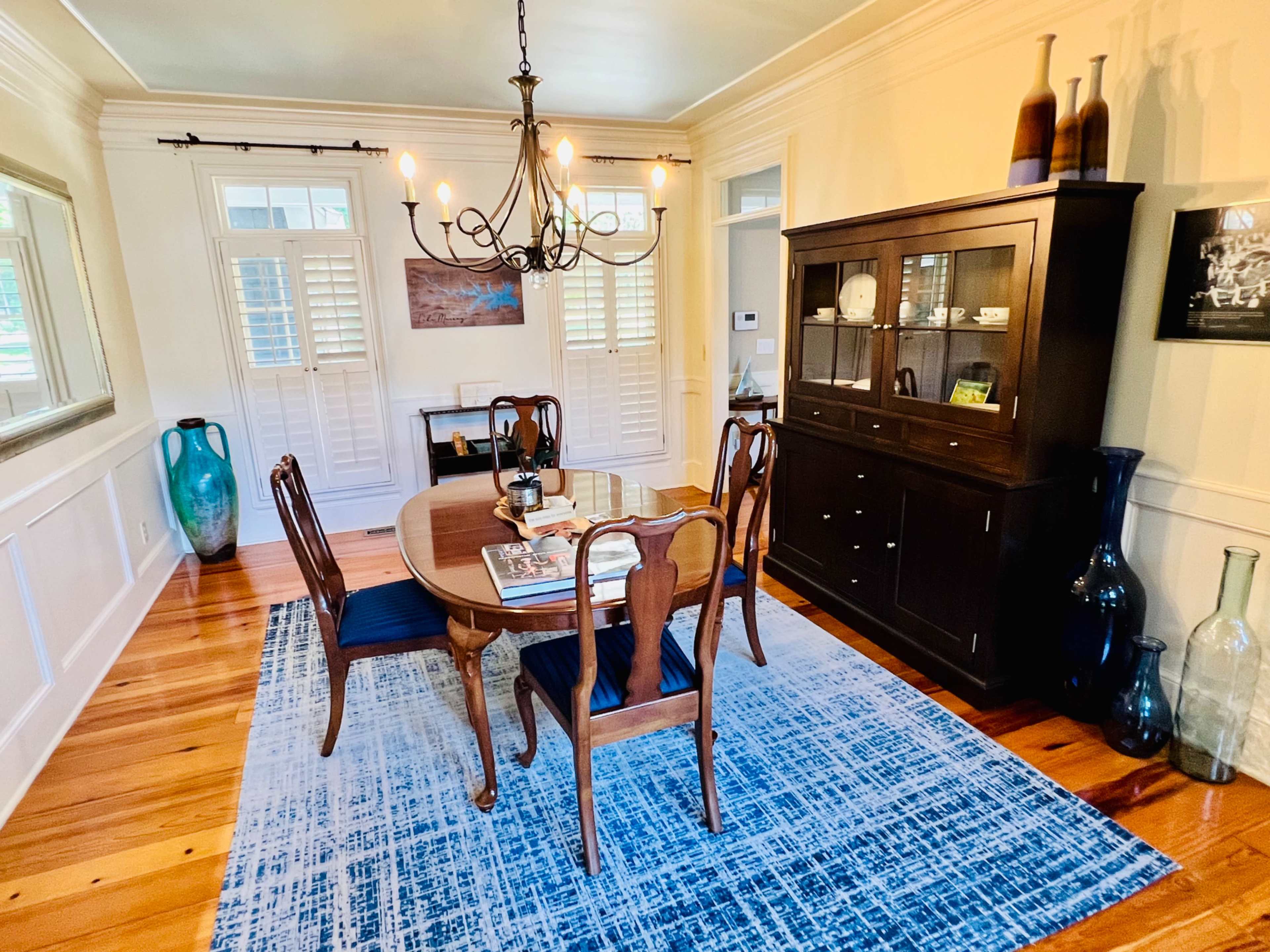 A dining room features a wooden table surrounded by four chairs, a chandelier overhead, and a cabinet displaying various items, with blue and green vases on the floor.