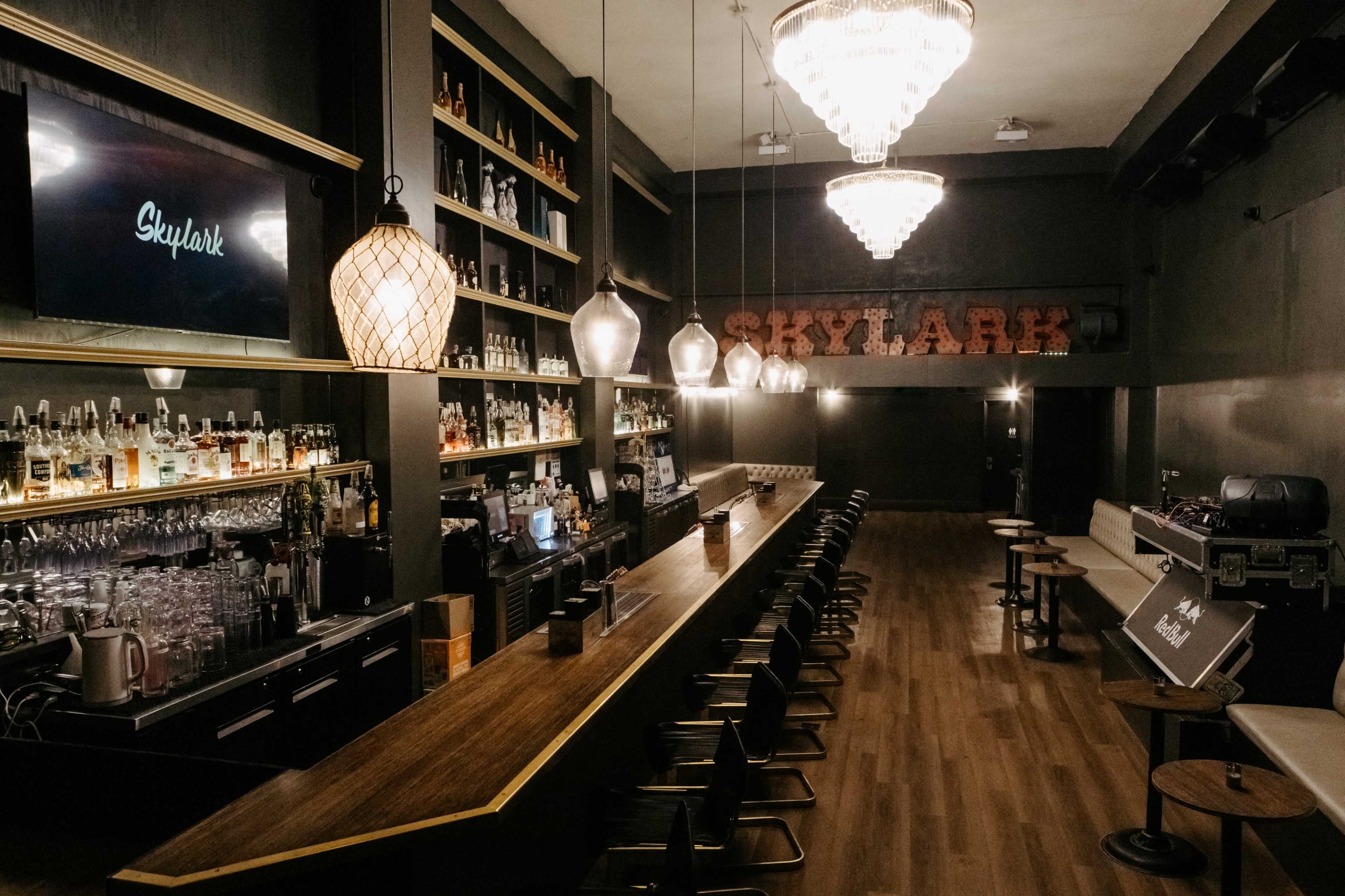 The image shows a dimly lit bar with a long wooden counter, shelves stocked with various bottles, and decorative pendant lights hanging from the ceiling.
