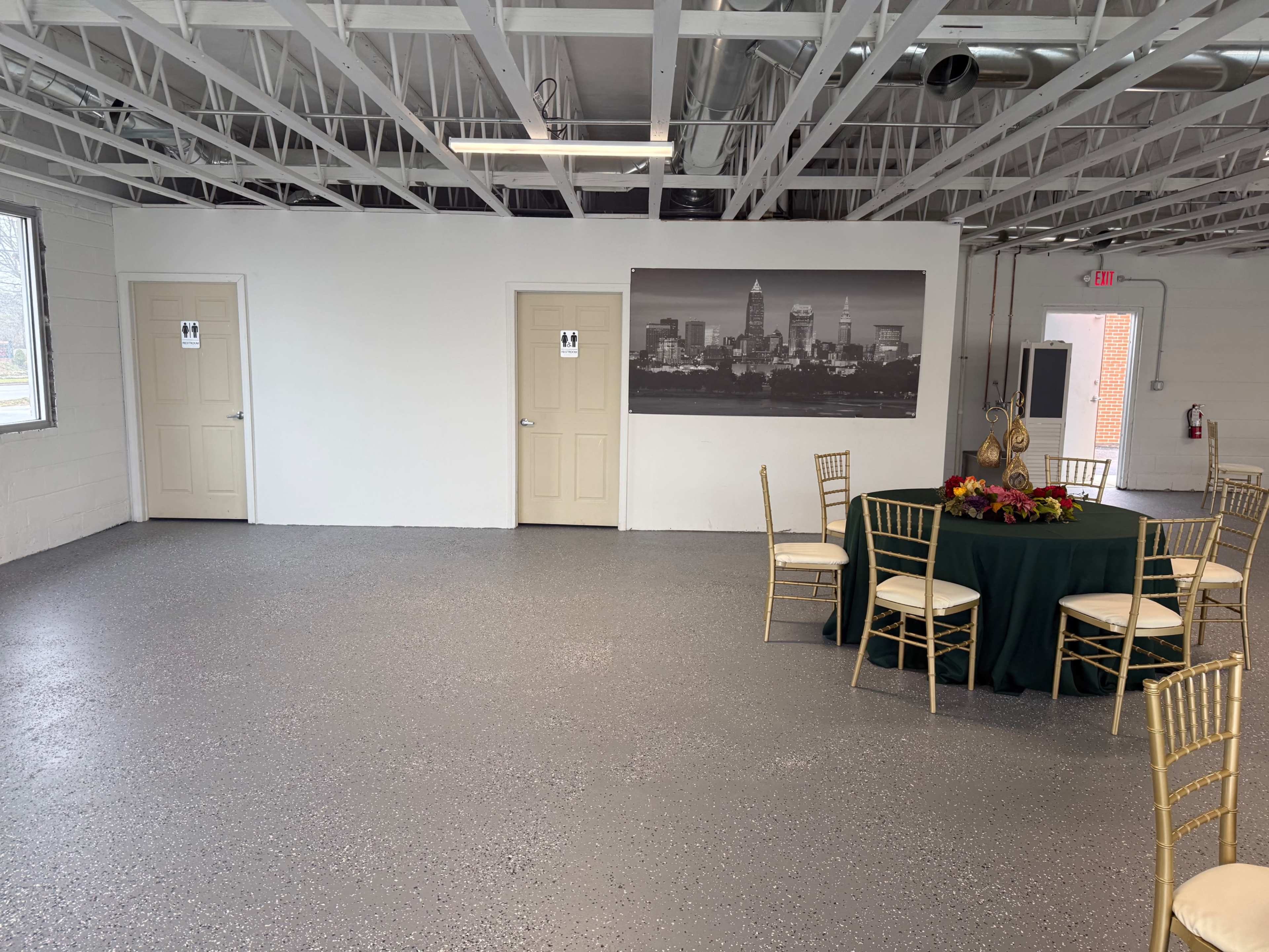 The image shows an empty room with two closed doors on one wall, a large photo of a city skyline, and a round table with chairs set up for an event.