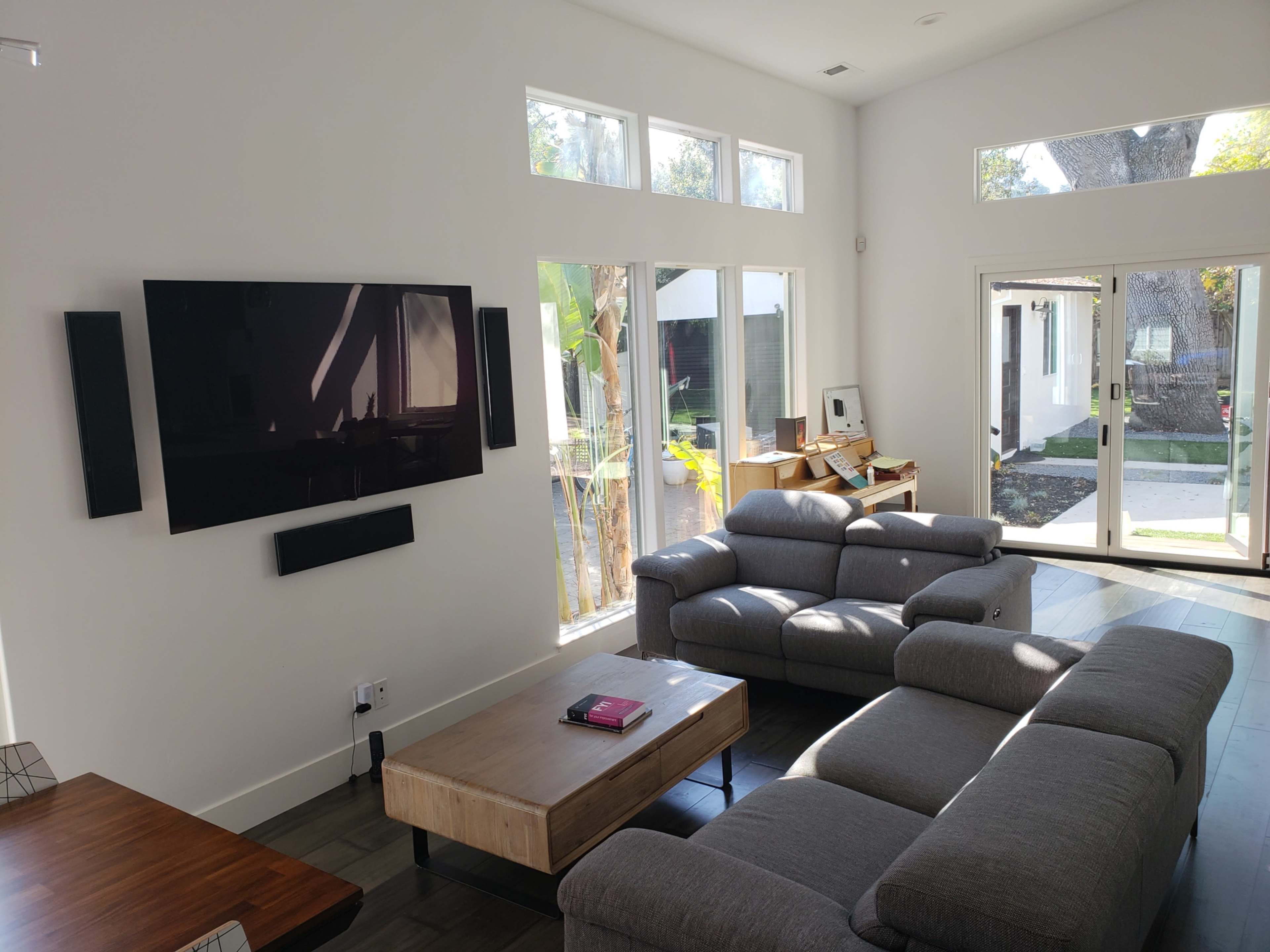 A modern living room features a television mounted on the wall, a gray sectional sofa, a wooden coffee table, and large windows allowing natural light.