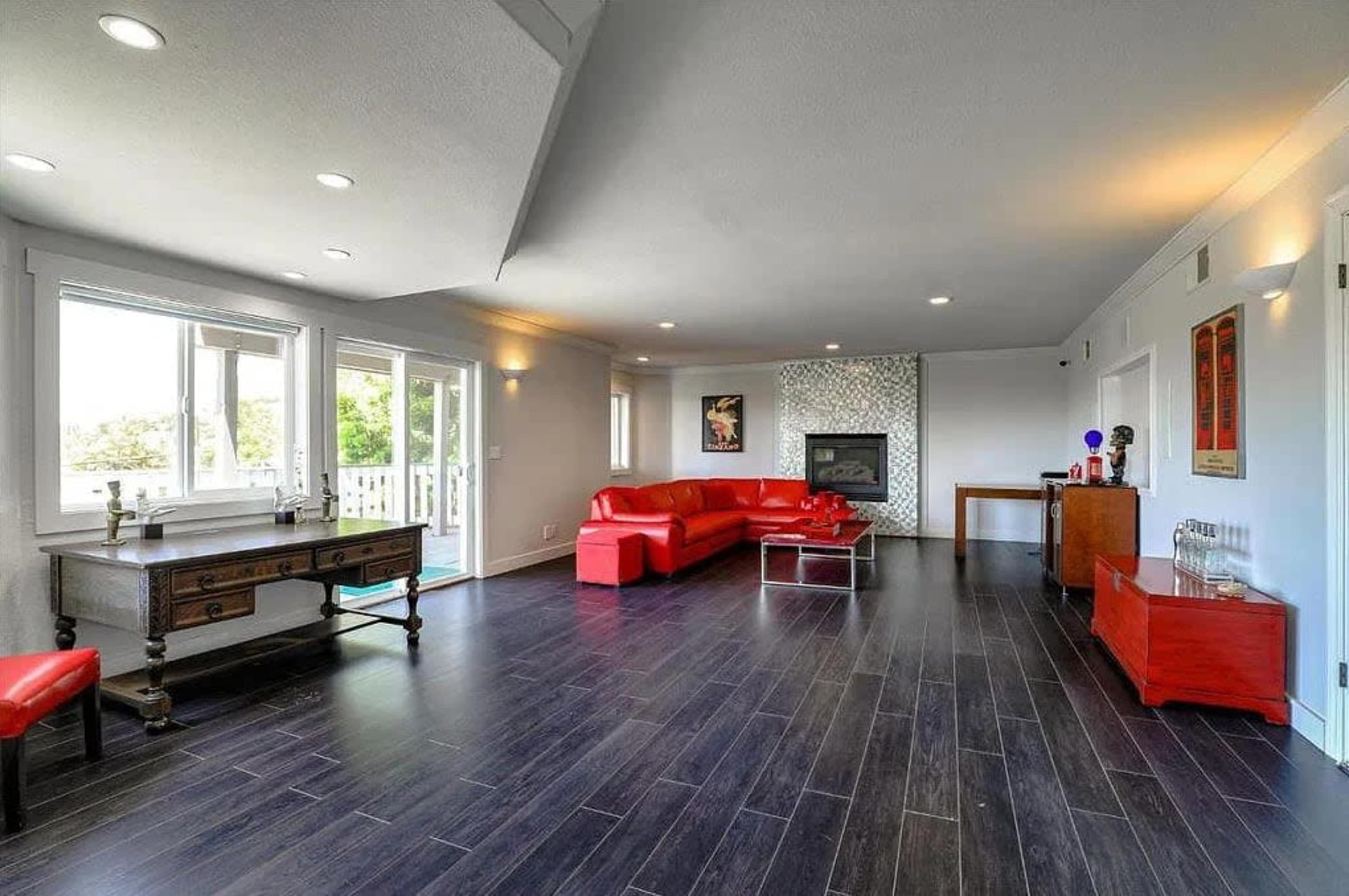 The image shows a modern living room featuring a red sectional sofa, a fireplace, large windows, and sleek dark flooring.