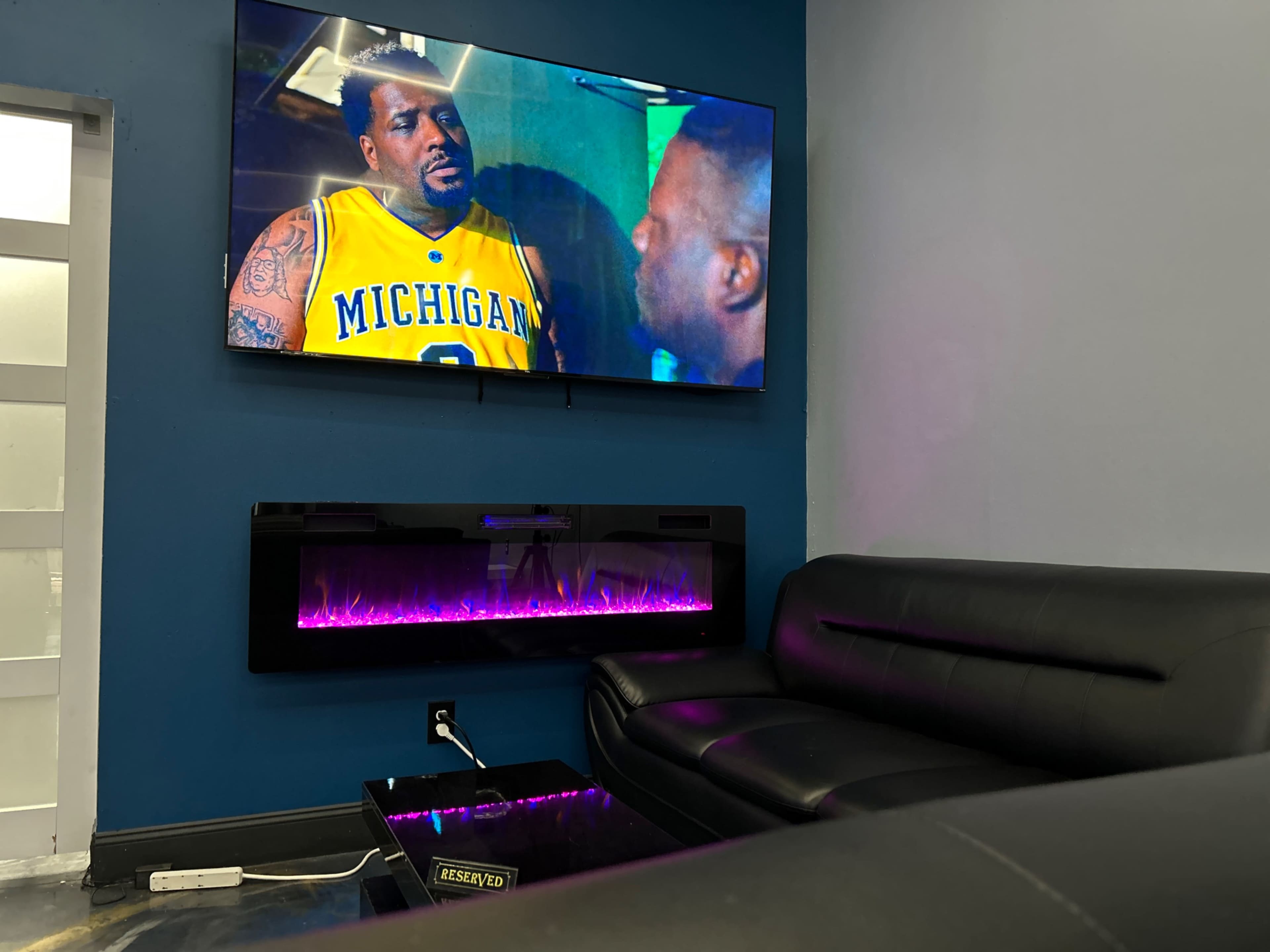 A black leather couch faces a wall-mounted television displaying a scene from a movie, while a modern electric fireplace with purple lighting is situated below the TV.