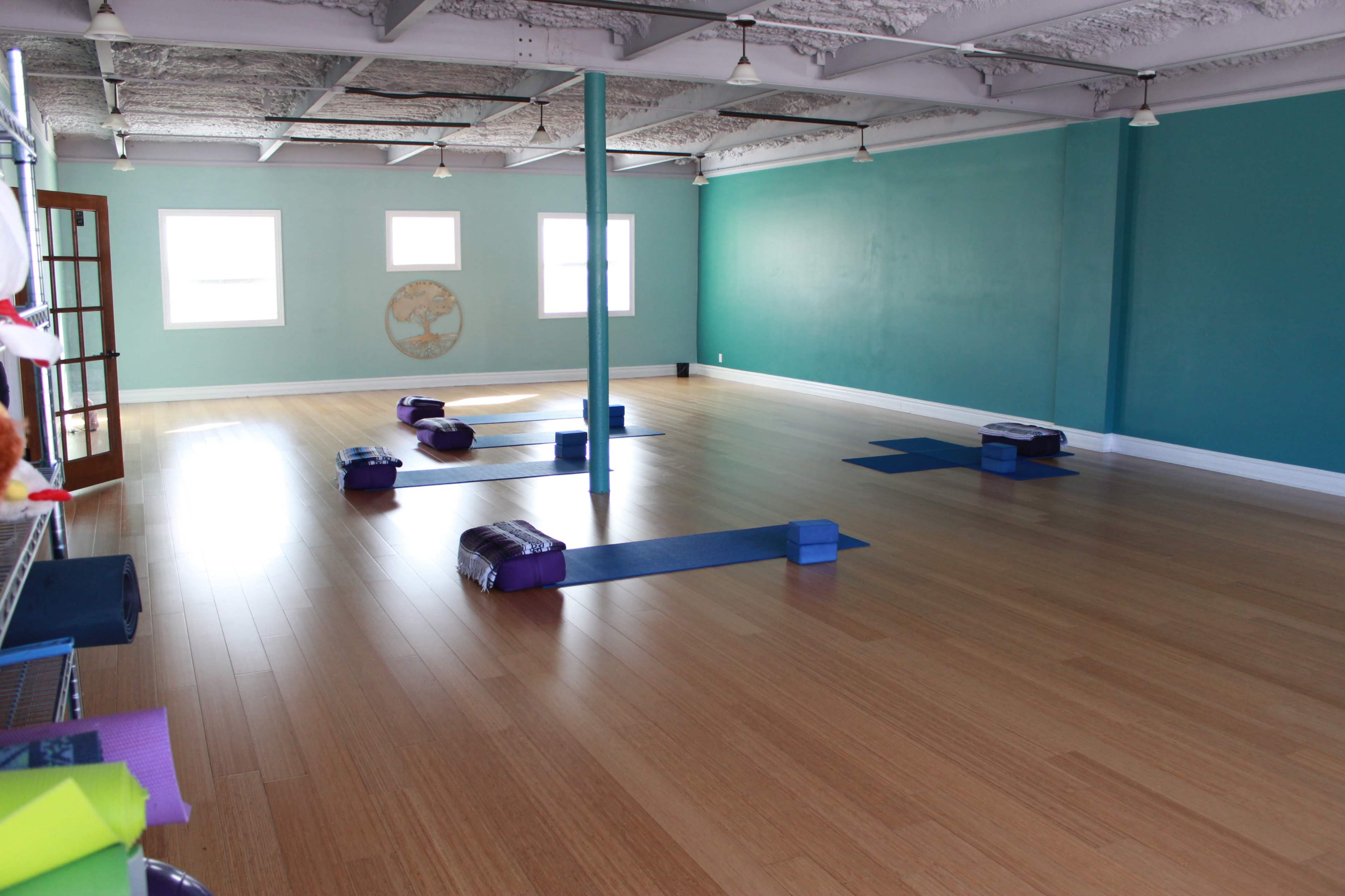 The image shows a spacious, empty yoga studio with wooden flooring, blue walls, and several yoga mats and blocks arranged on the floor.