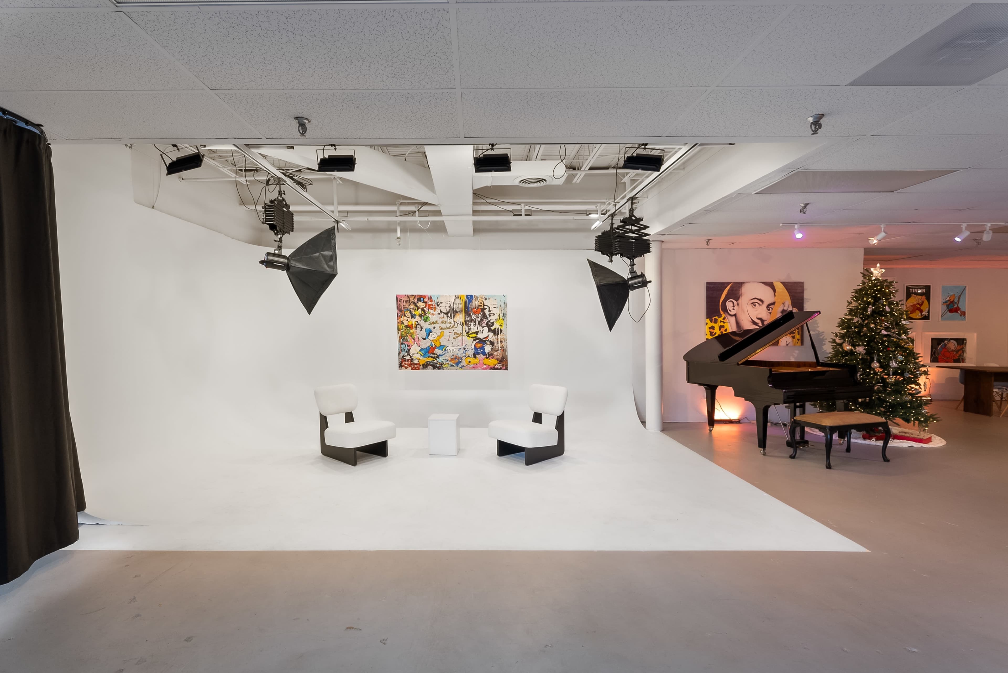 The image shows a studio space featuring a white backdrop, two black and white chairs, a small white table, a colorful artwork on the wall, a grand piano, a Christmas tree, and various paintings displayed in the background.