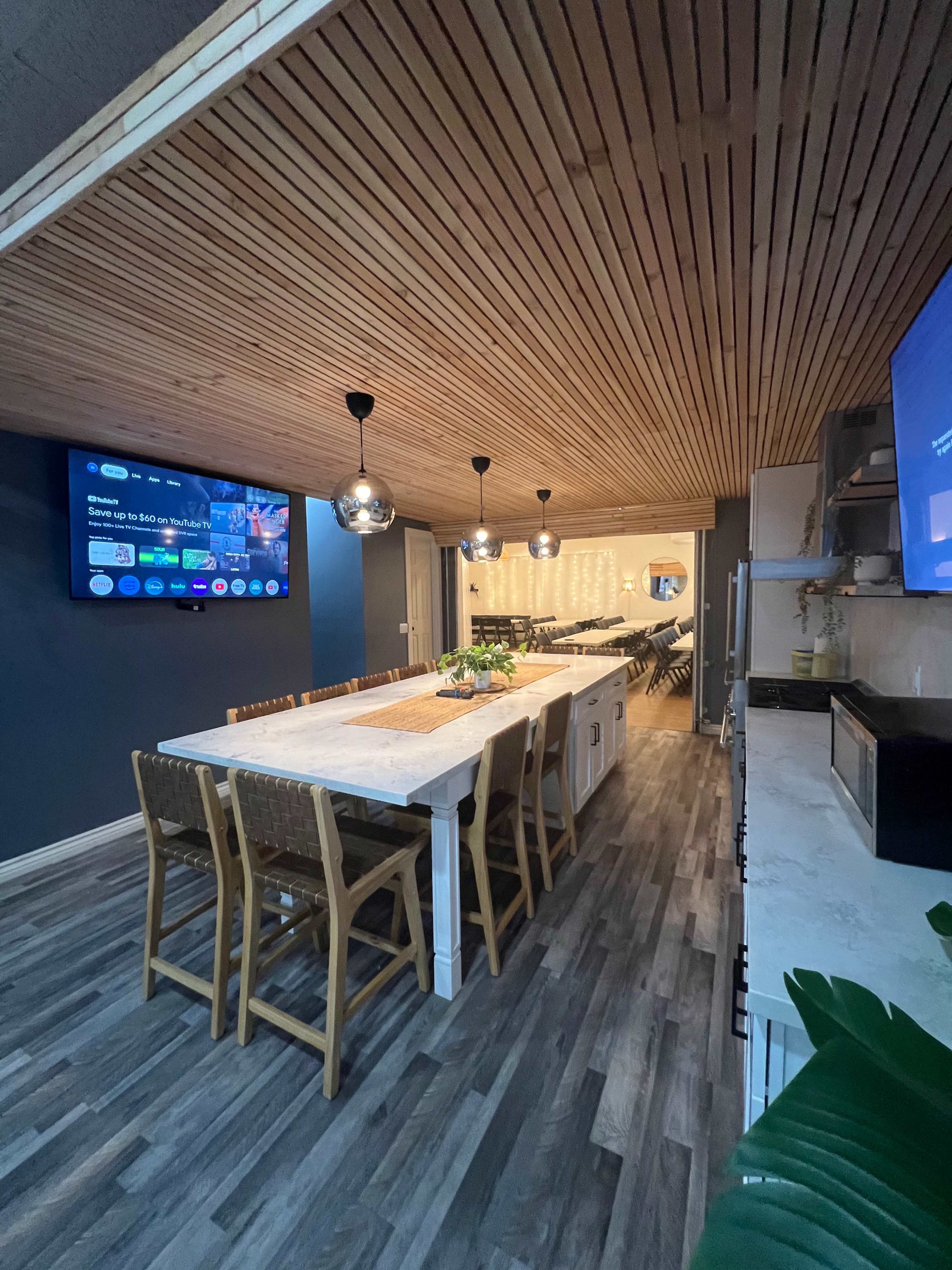 A modern kitchen features a long dining table with seating, wooden ceiling panels, and two wall-mounted televisions.
