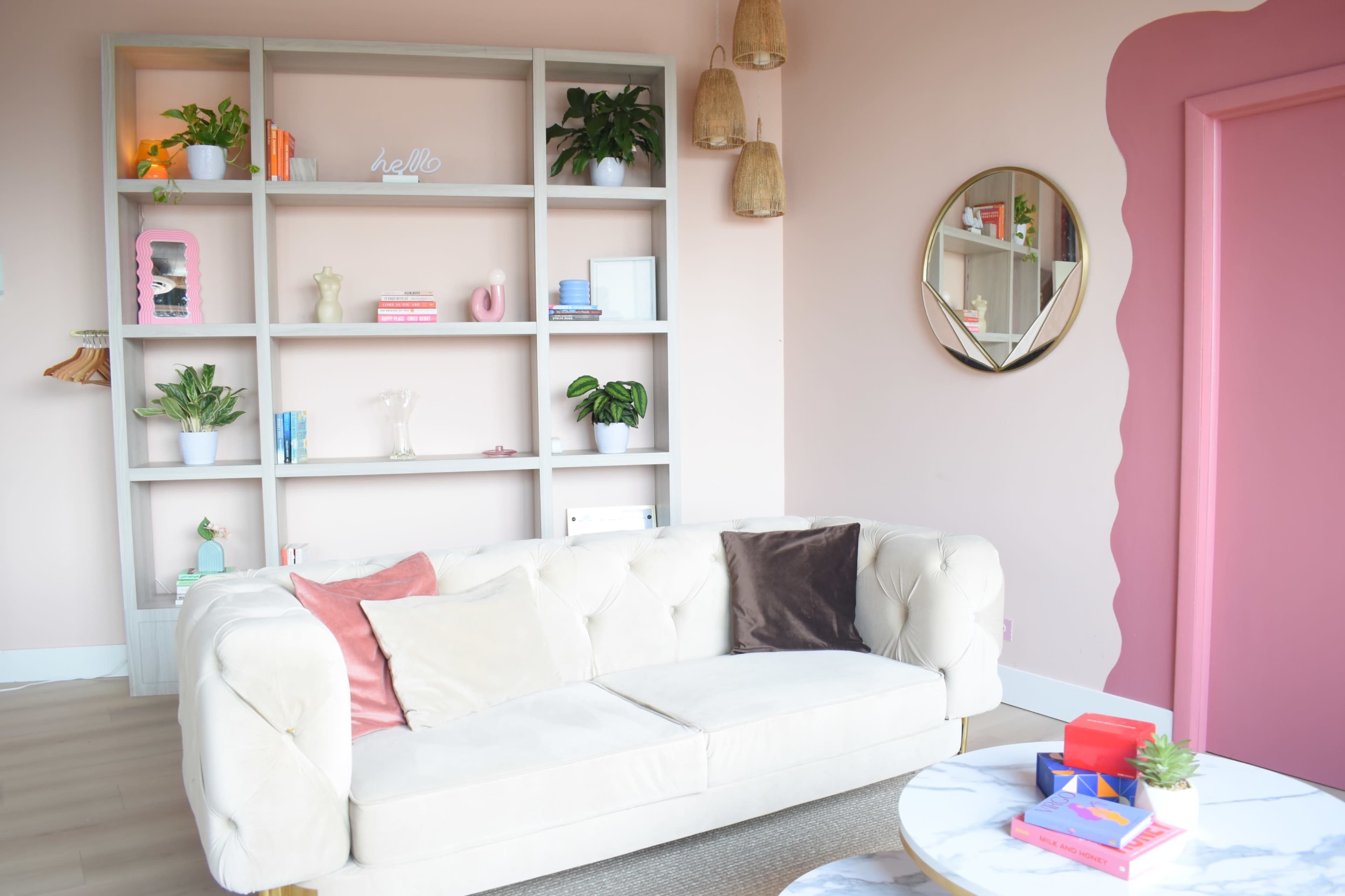 A light-colored couch with decorative pillows is positioned in front of a pink wall featuring a shelf filled with books and plants.