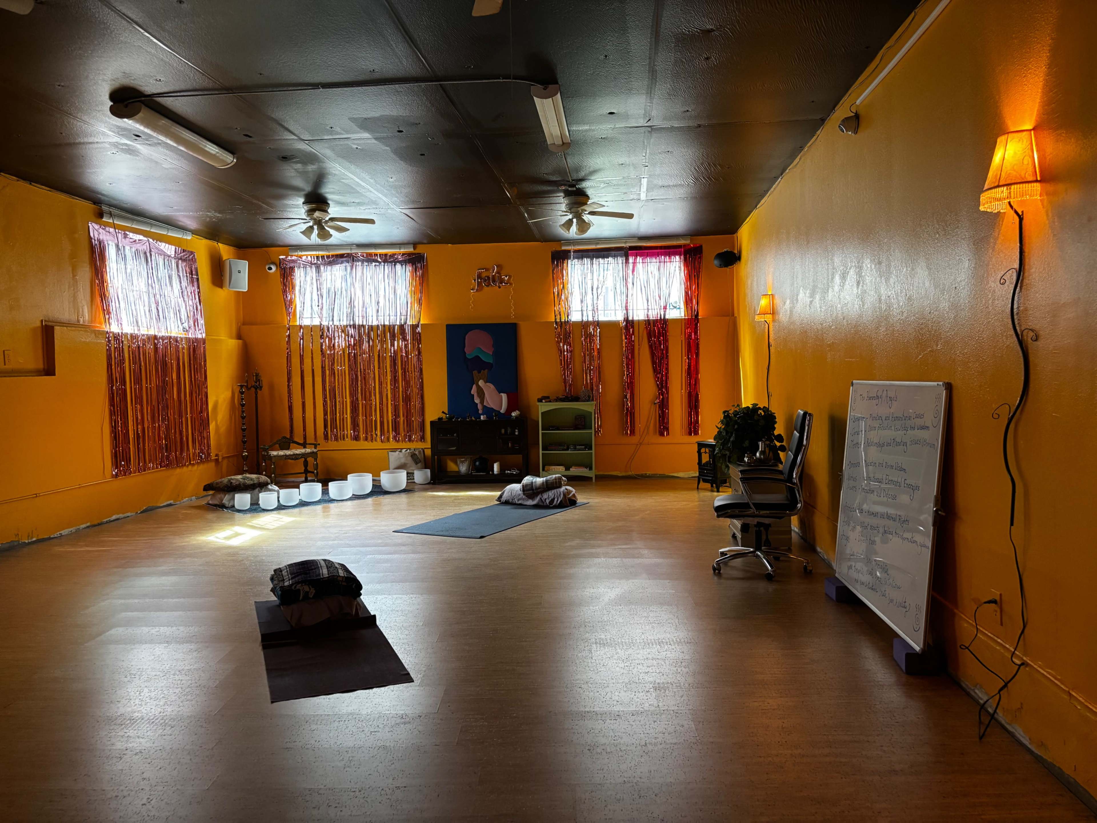 The image depicts a brightly colored yoga studio with orange walls, floor cushions, yoga mats, and a crystal sound bath setup.