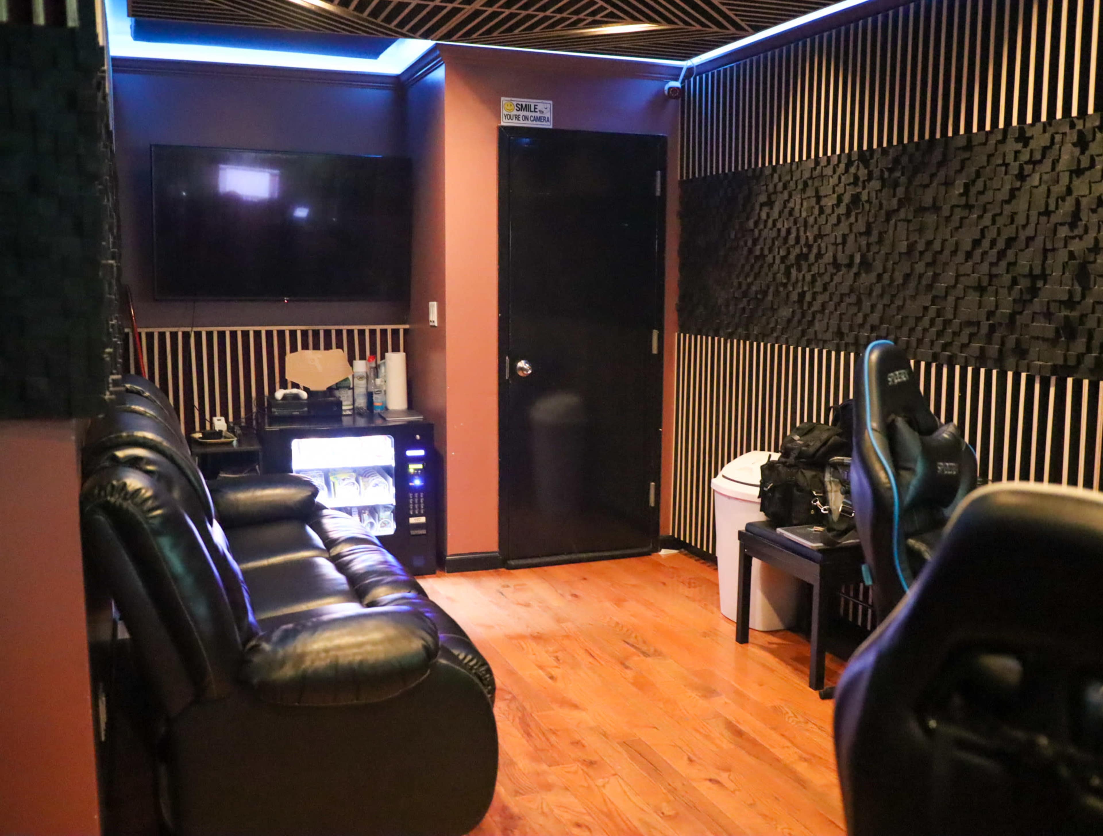A room with leather seating, a television mounted on the wall, and a door leading to another area, featuring wooden flooring and soundproof wall panels.