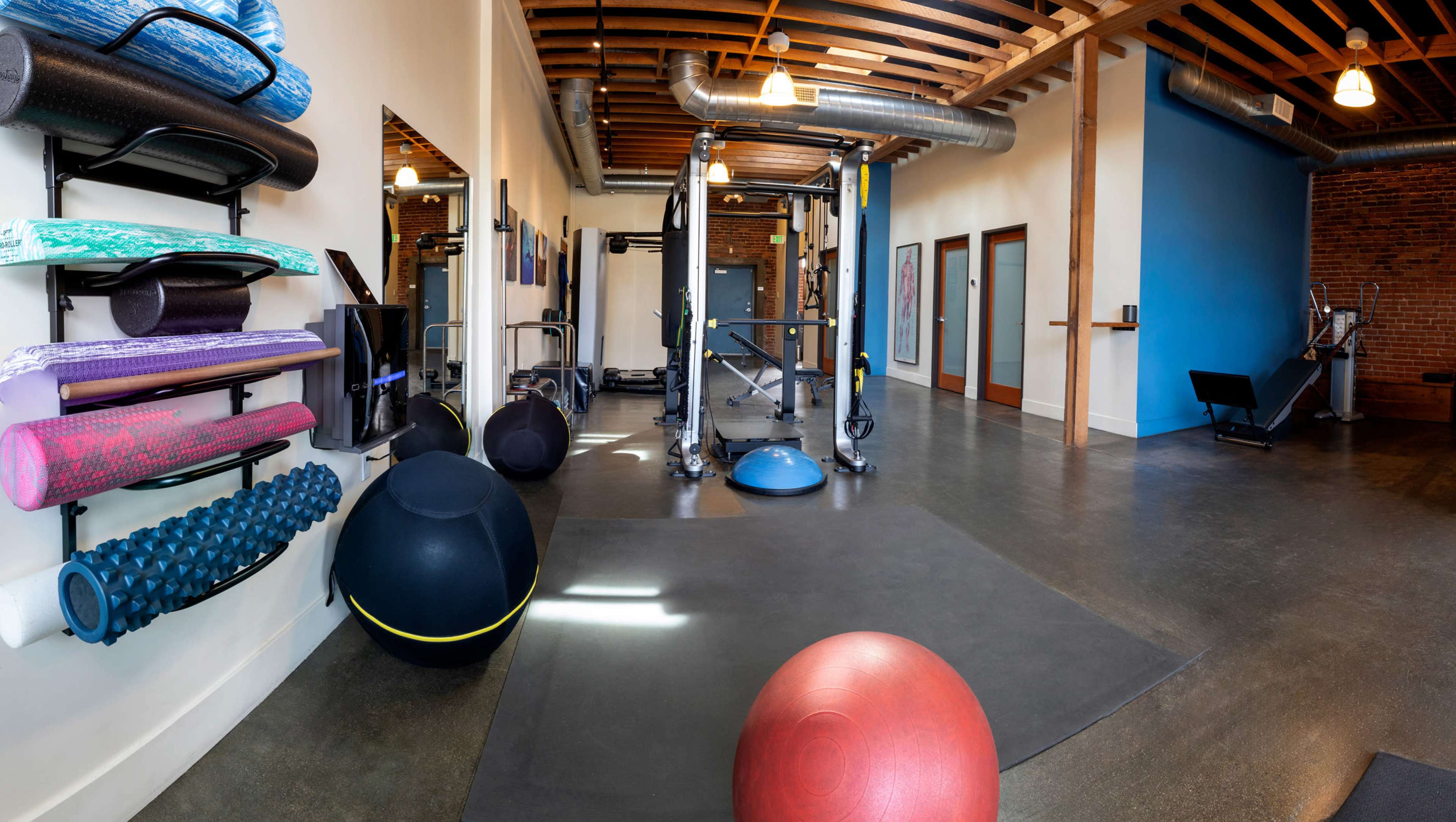A fitness studio with various exercise equipment, including yoga mats, stability balls, and a multi-functional gym machine, under exposed wooden beams and soft lighting.
