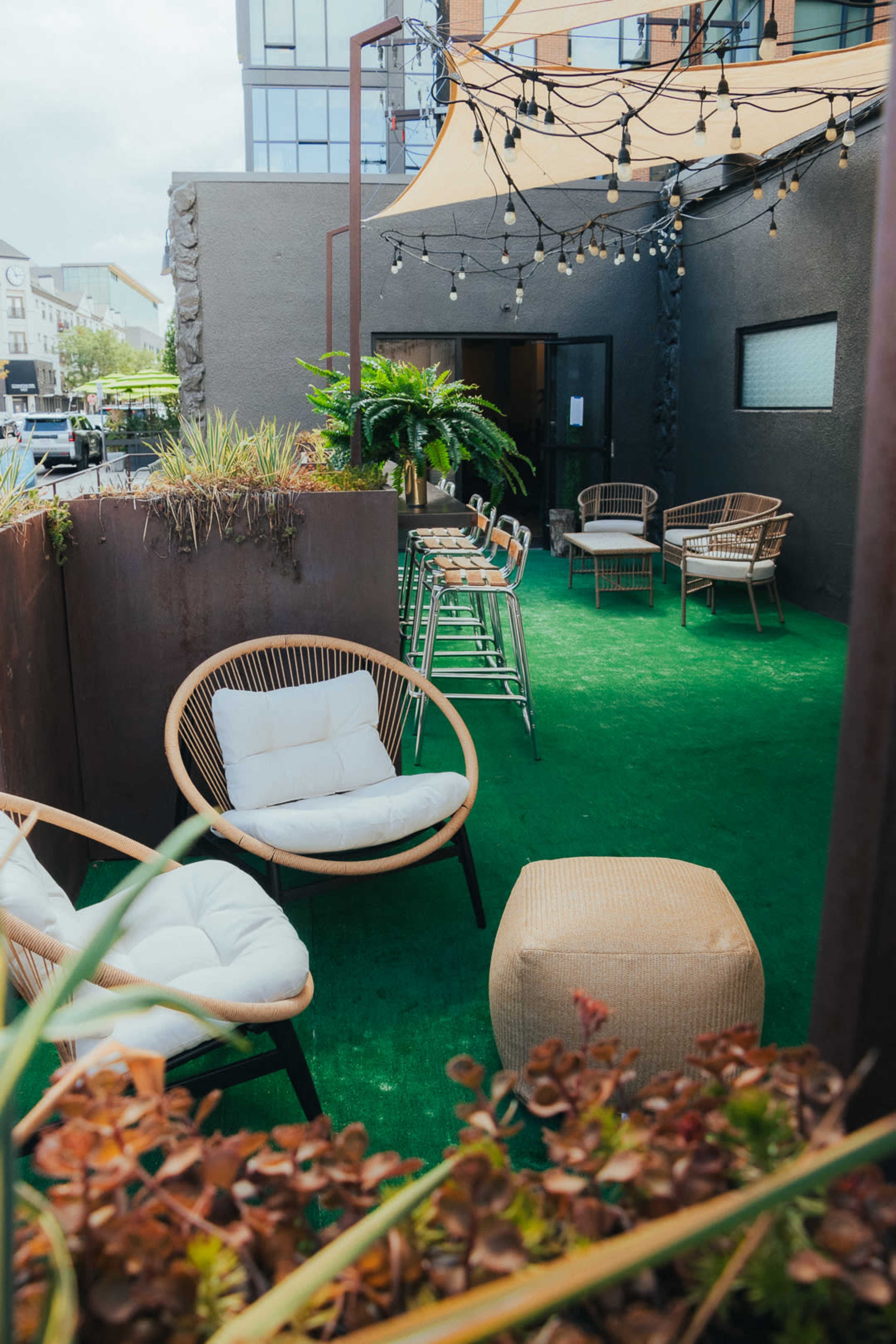 The image shows an outdoor seating area with several chairs and tables on green grass-like turf, surrounded by plants and string lights.