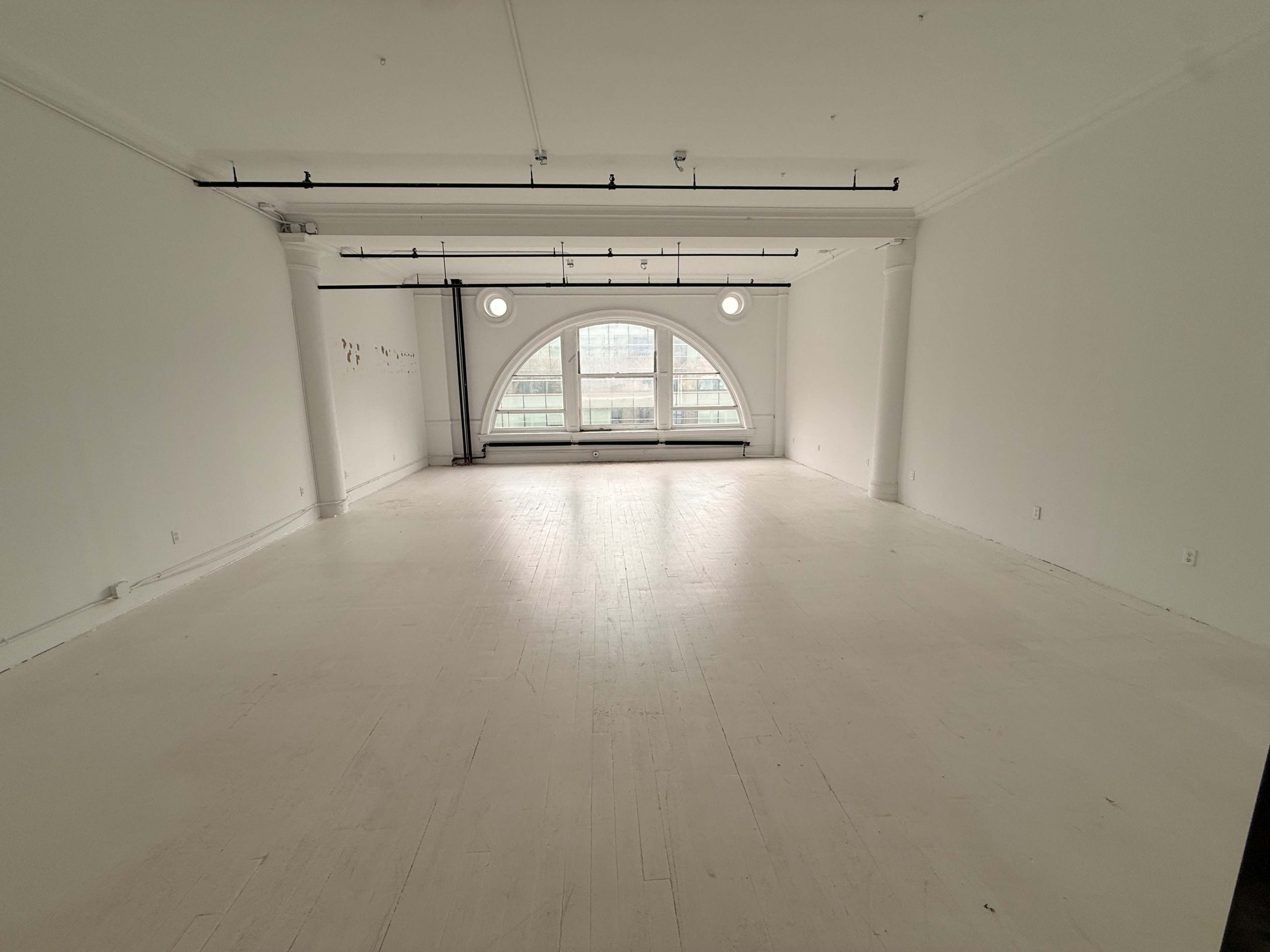 The image shows a spacious, empty room with white walls, a light-colored wooden floor, and large arched windows providing natural light.