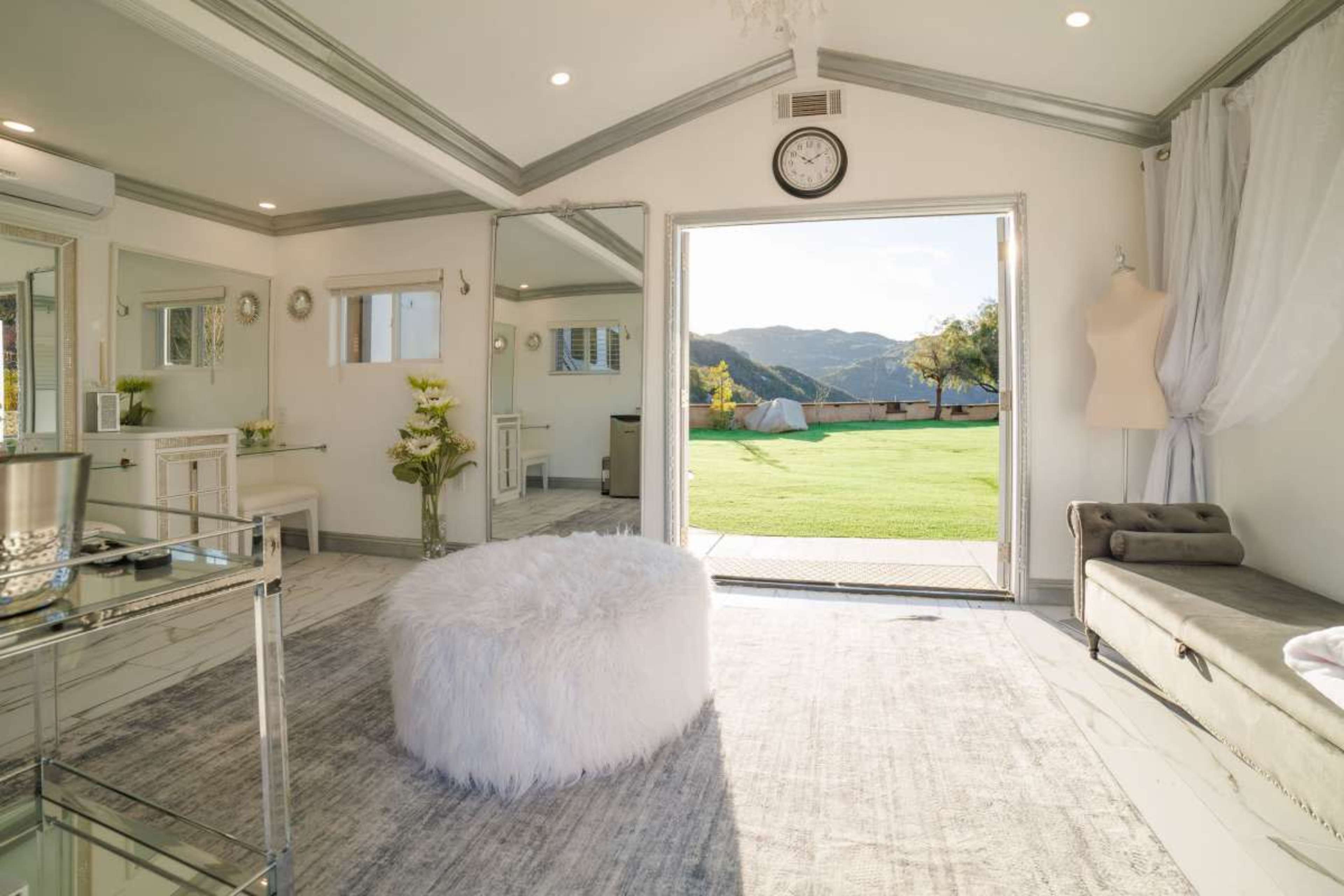 The image shows a bright, modern room featuring a fluffy white ottoman, a large mirror, and a view of a grassy outdoor area through an open door.