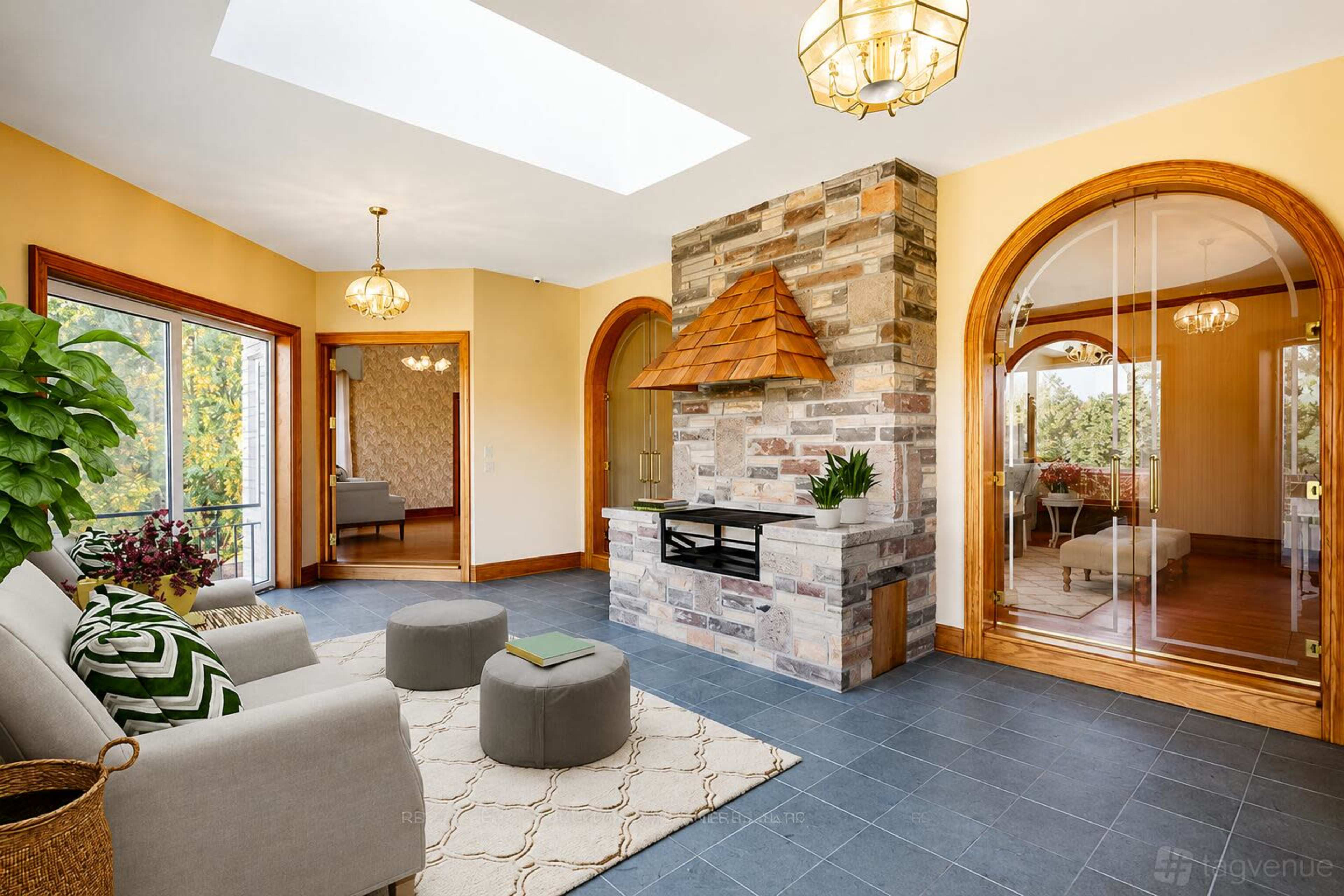 The image shows a well-lit sunroom featuring a stone fireplace, two circular ottomans, and large windows overlooking greenery.