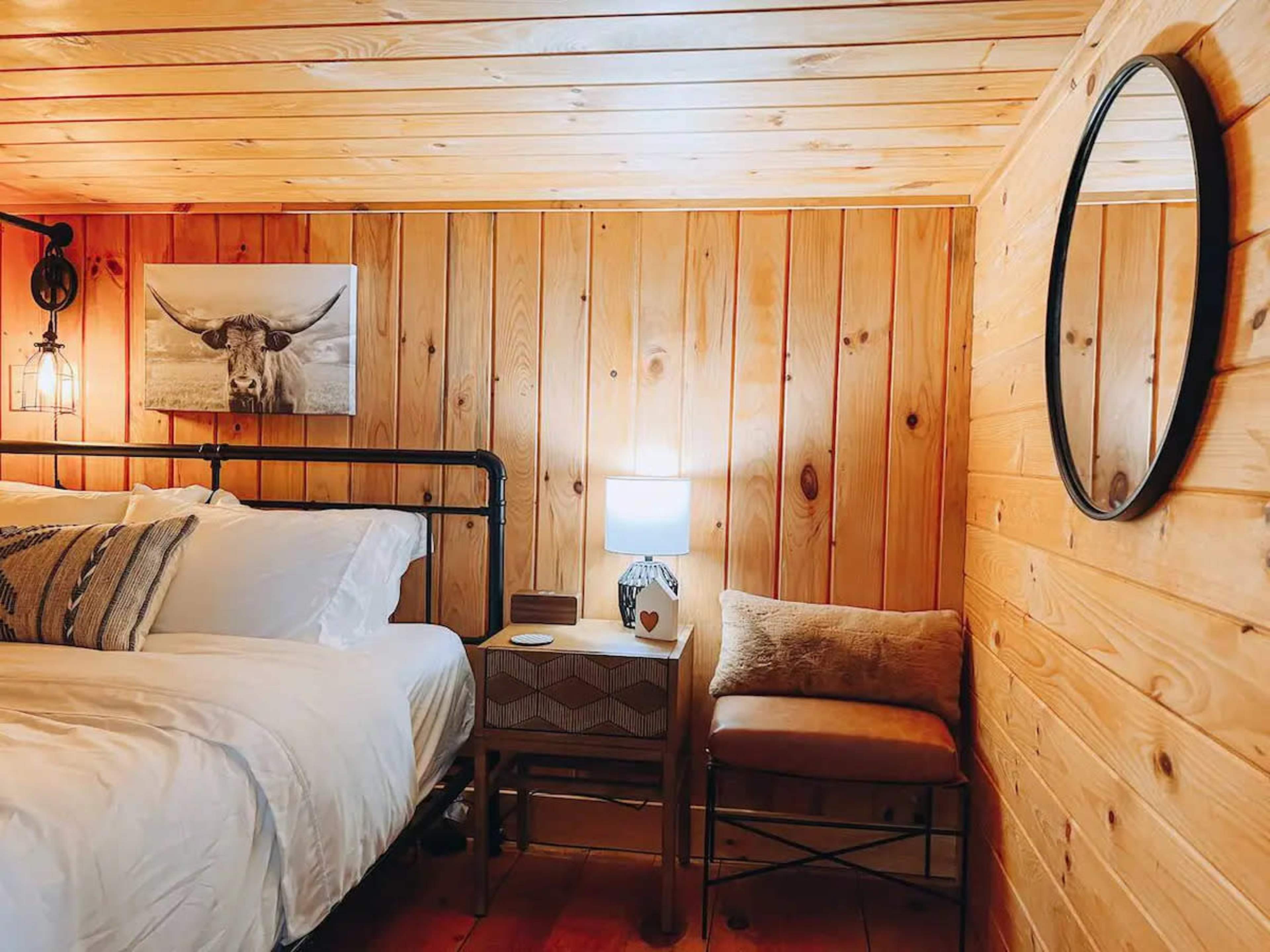 A cozy bedroom with wooden walls, a bed with white bedding, a small bedside table with a lamp, and a mirror on the wall.