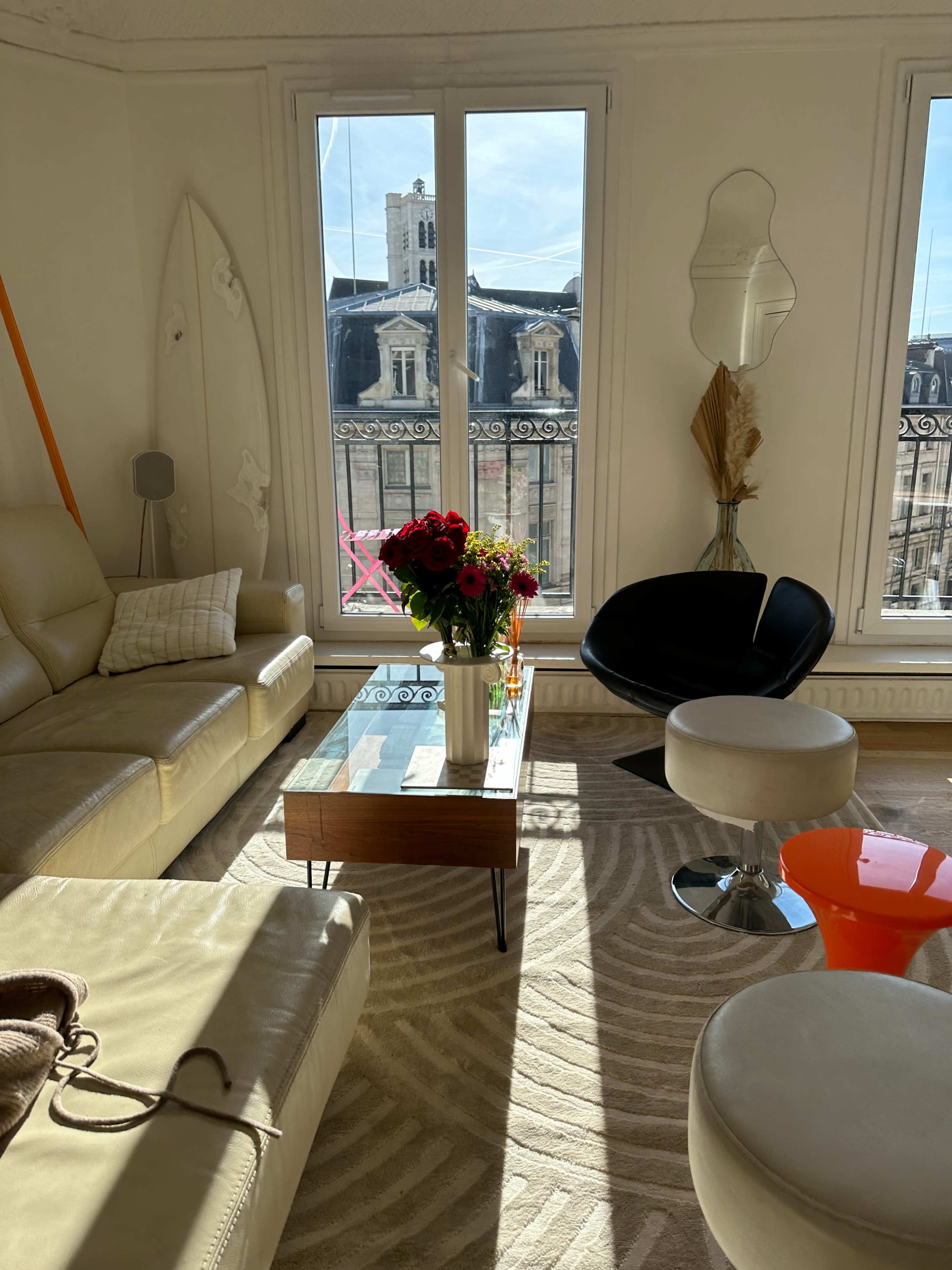 The image shows a modern living room with a beige leather sofa, a glass coffee table, and a variety of decorative elements, all lit by natural light streaming through large windows.
